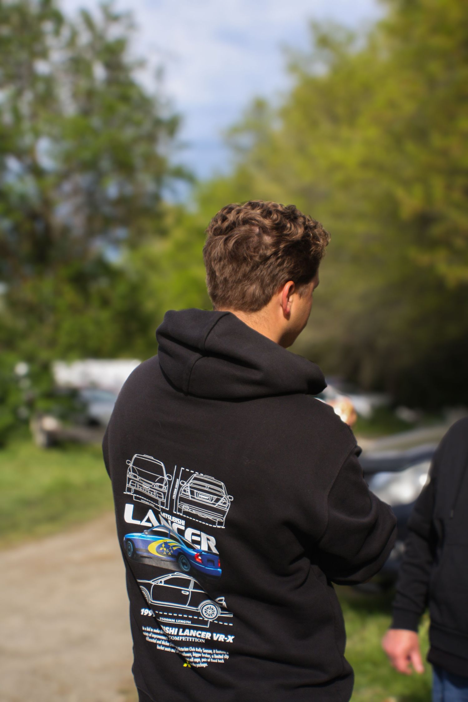 Person wearing black hoodie with automotive design at outdoor racing event.