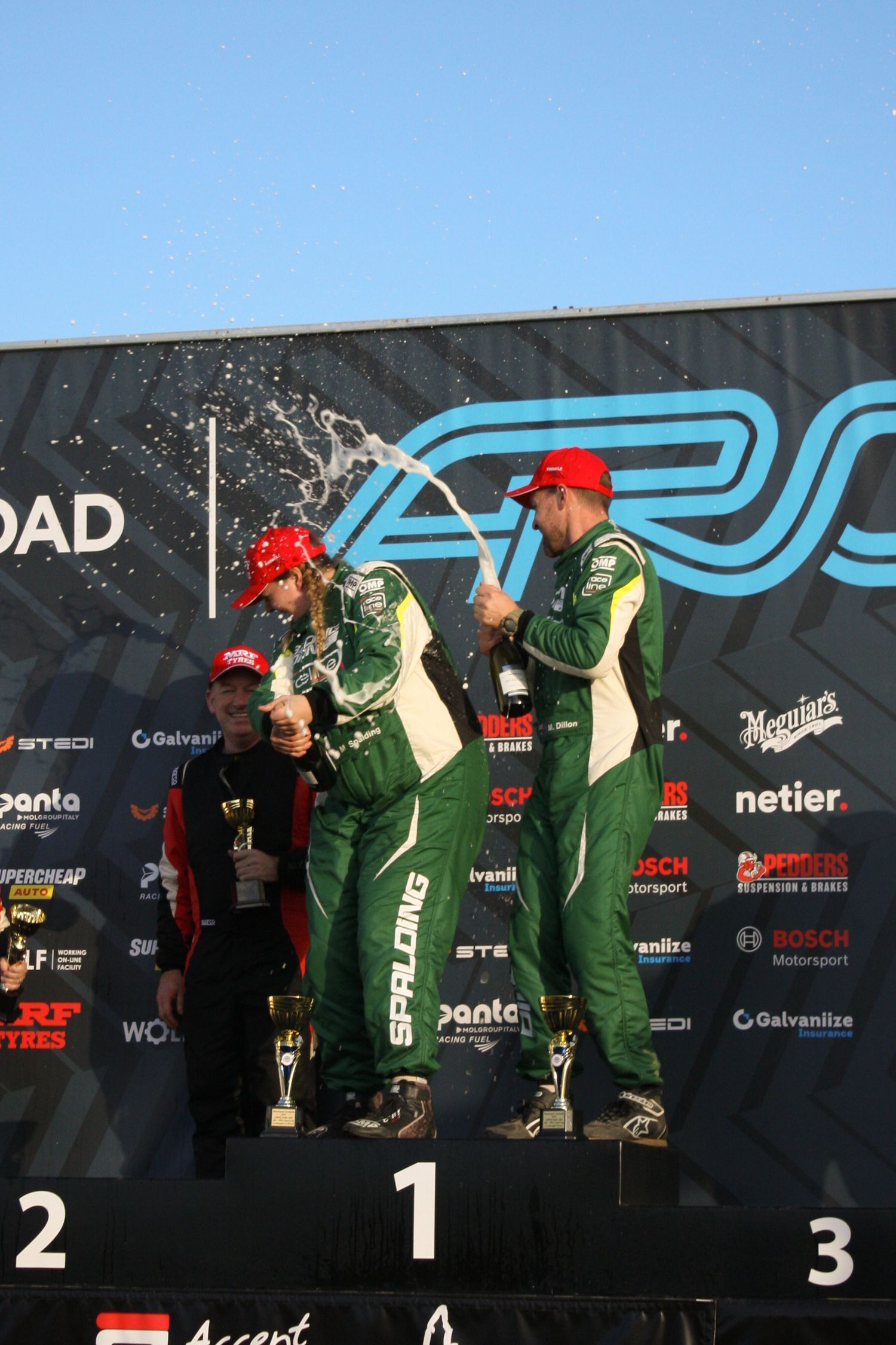 Racing team members celebrate on a podium with champagne spray.