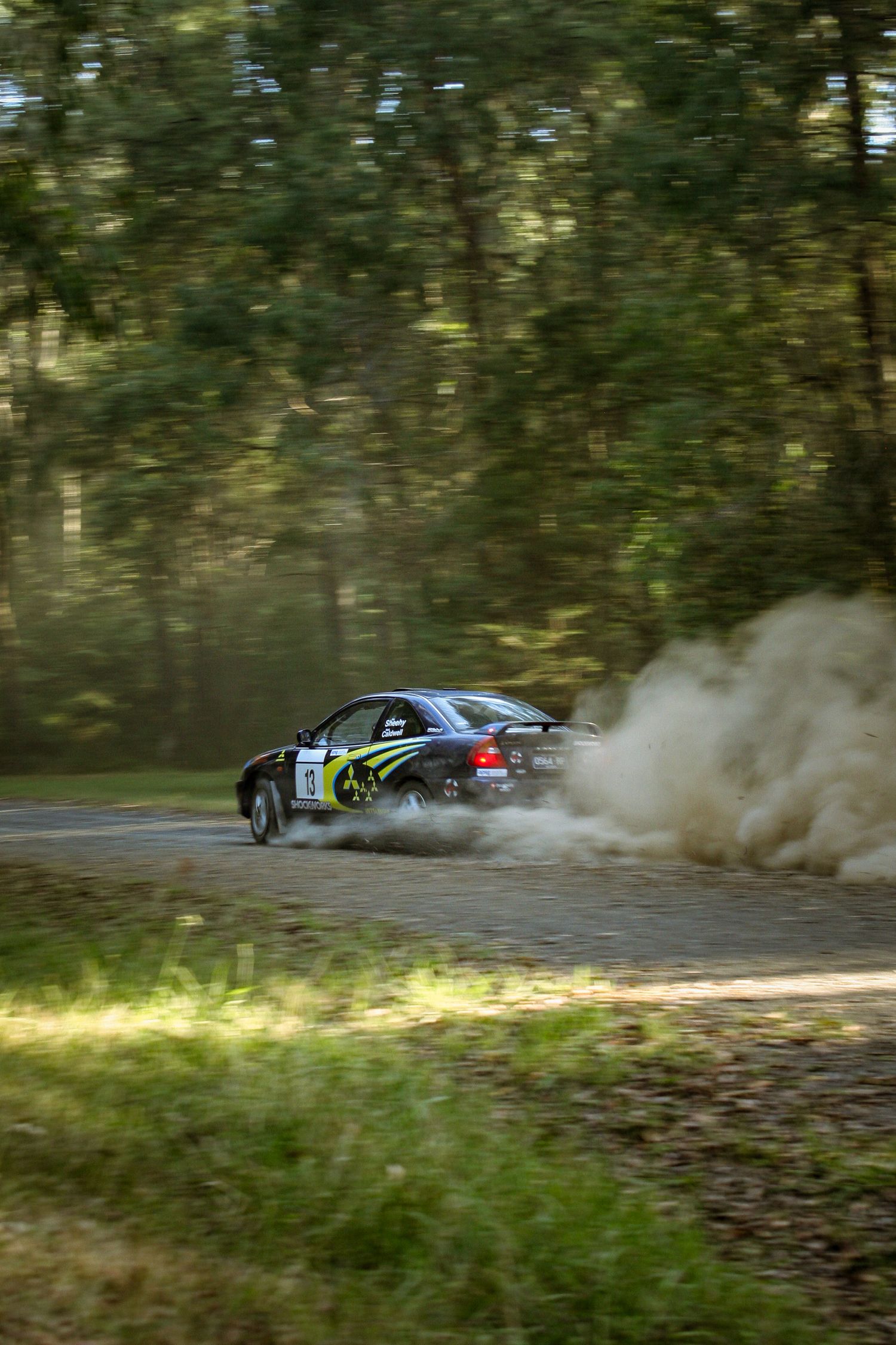 Blue rally car drifting through a dusty forest turn during competition.