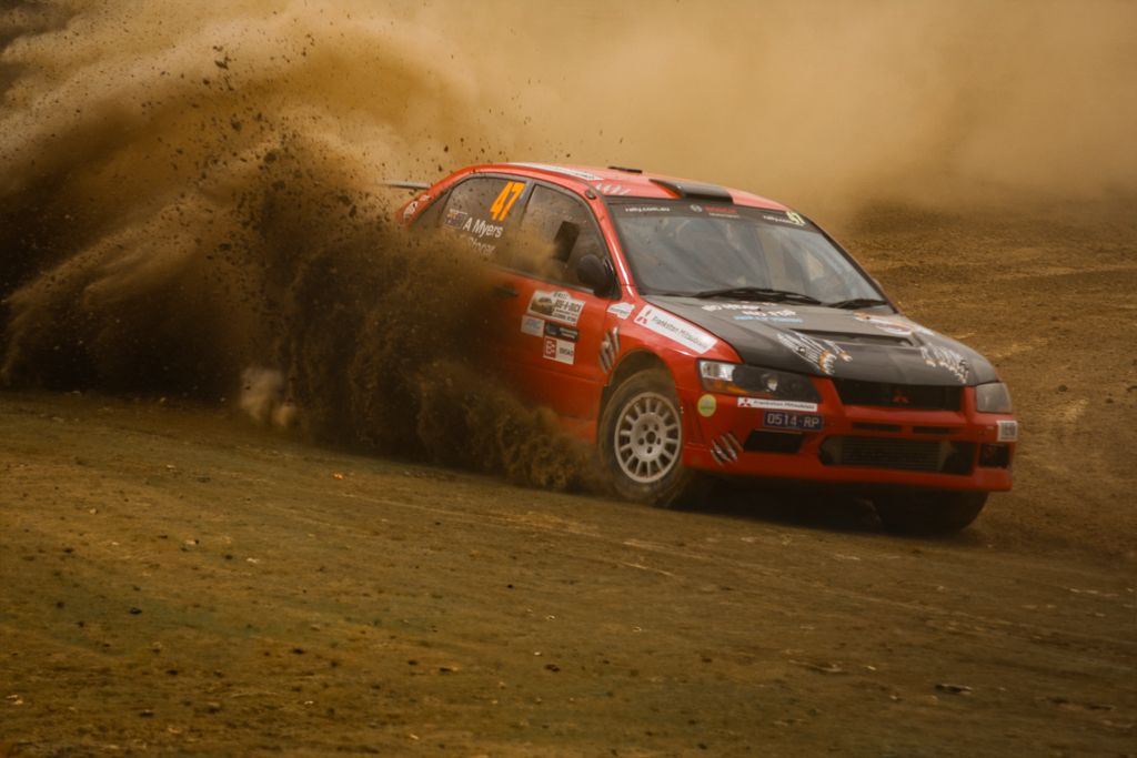 Red rally car racing through dirt track creating dramatic dust clouds at sunset.