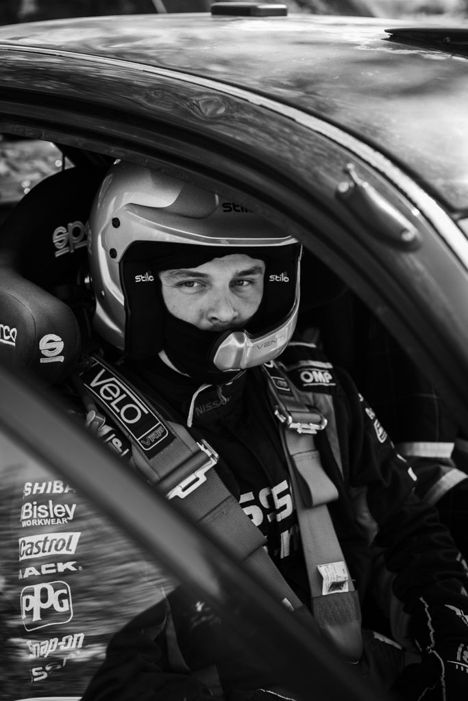 Racing driver in full safety gear and helmet sits in competition vehicle cockpit.