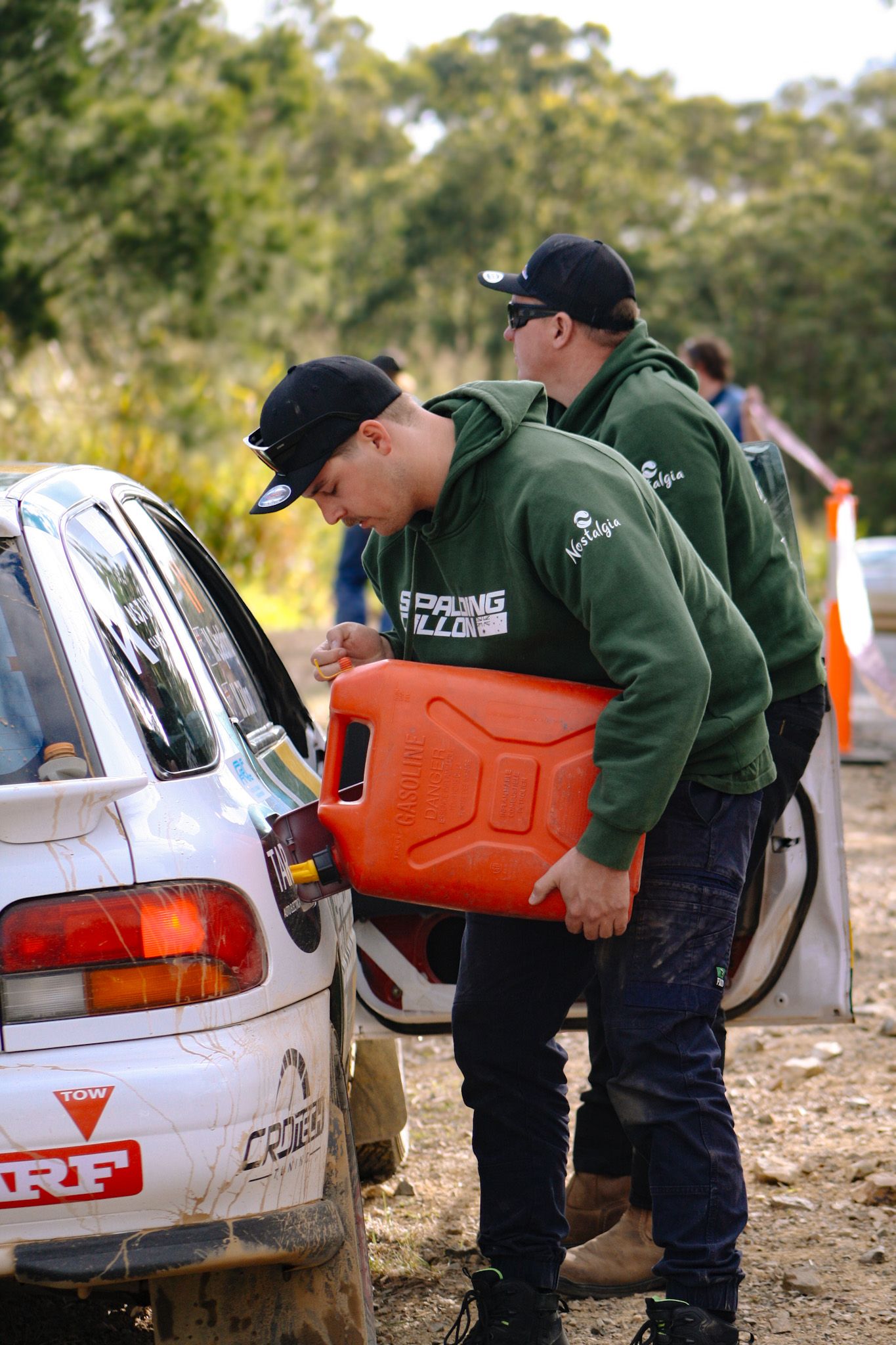 Team members refueling a white rally car with orange fuel containers.