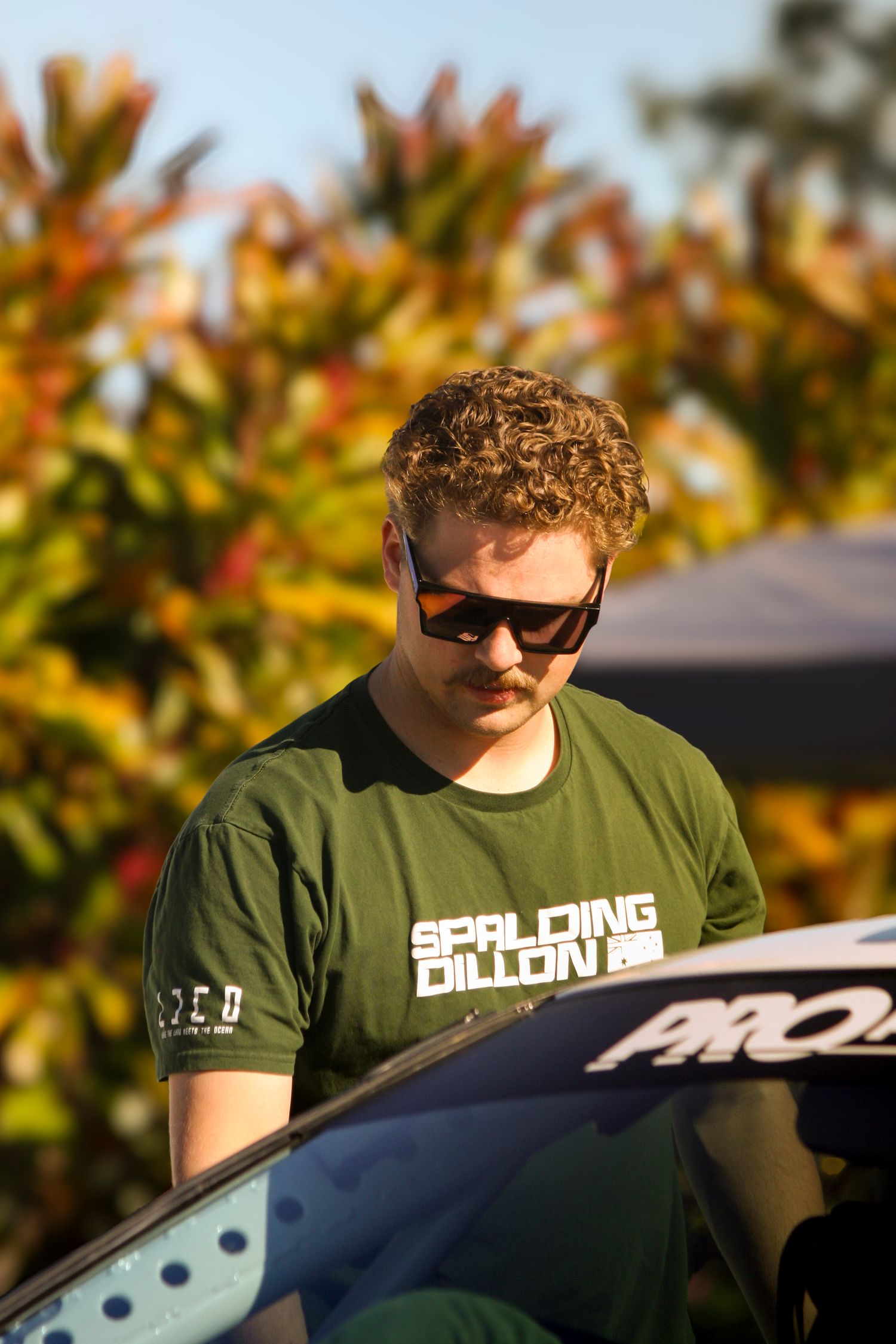 Rally team member wearing a green t-shirt stands near a race car in autumn setting.
