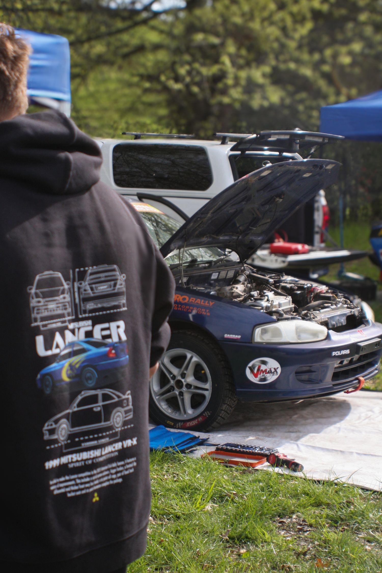 Rally car maintenance under blue tent with Lancer hoodie in foreground.