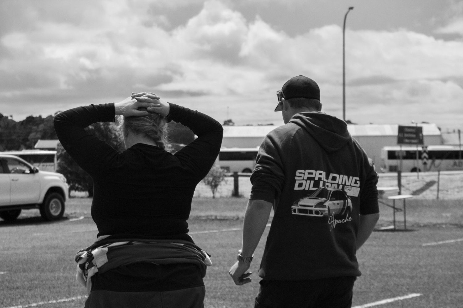 Two people in racing gear stand in a parking area during an overcast day.