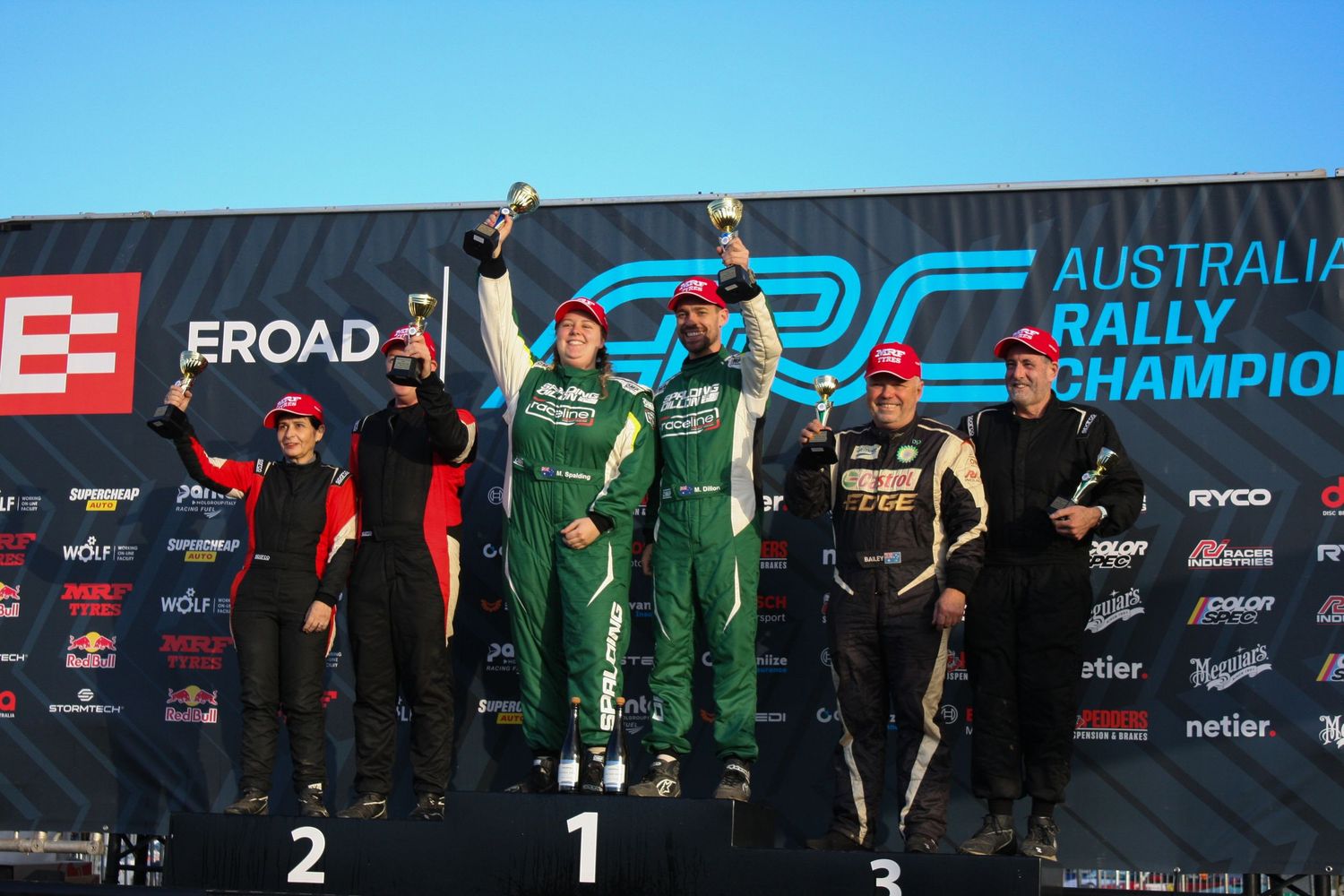 Winners celebrate on a motorsport podium with trophies under a branded backdrop.