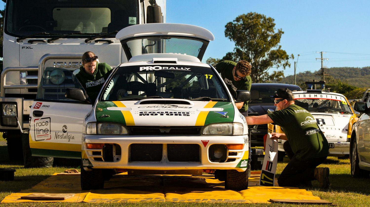 Racing team works on a white rally car in an outdoor service area.