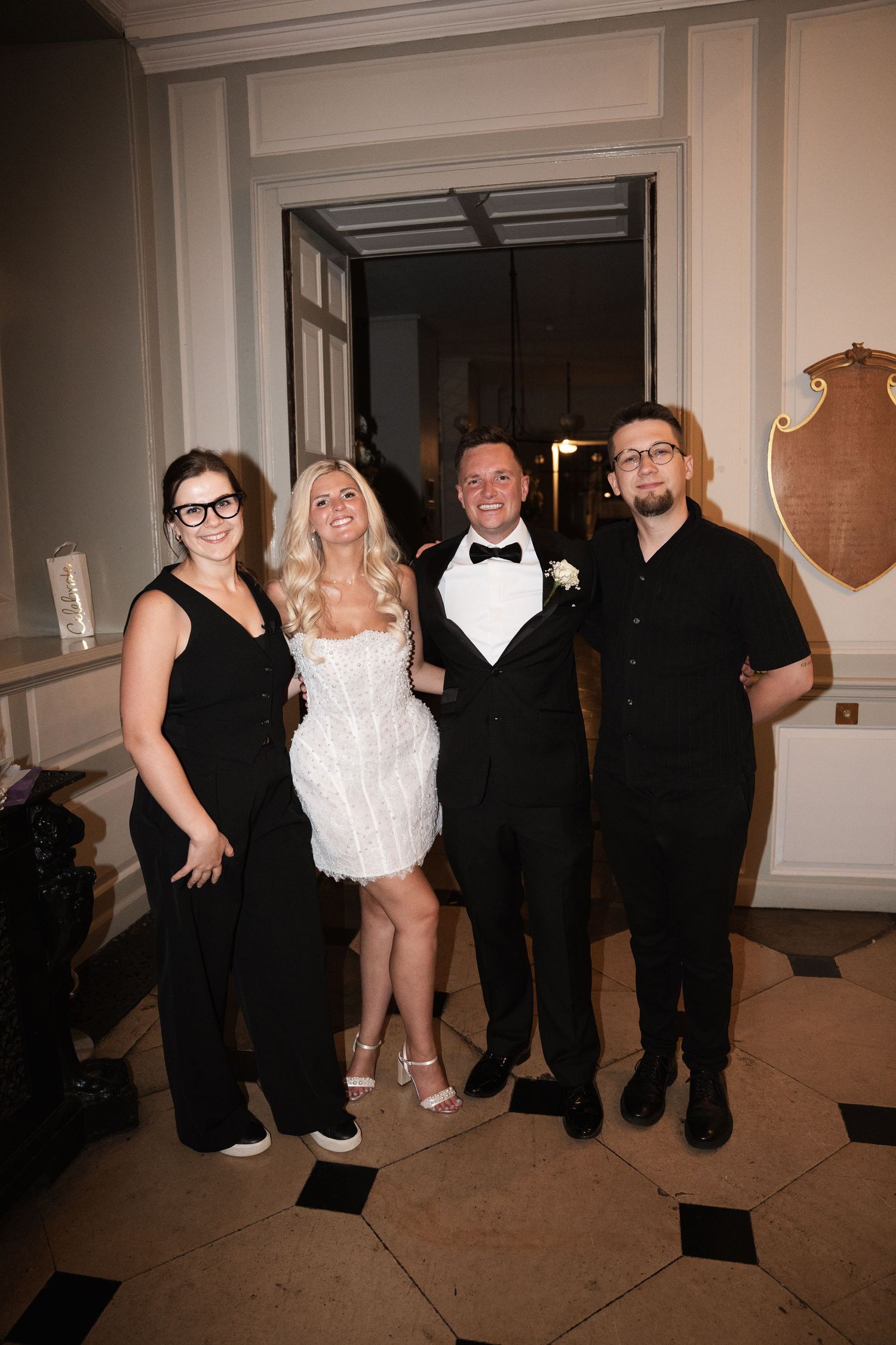 Four people pose together at a formal event, with one wearing a white dress and others in black attire.