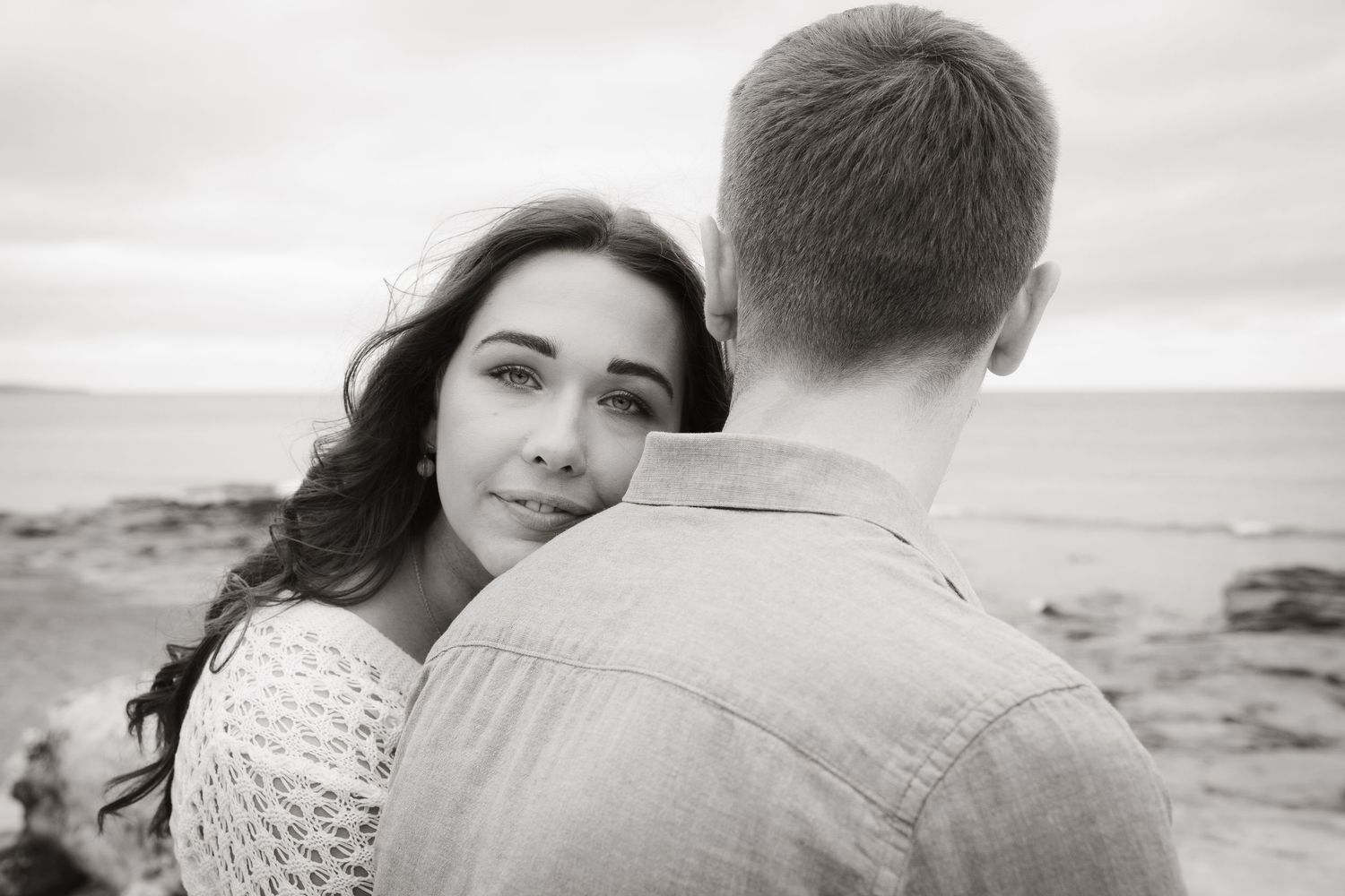 A romantic black and white portrait of a couple embracing at the beach with an overcast sky in the background.