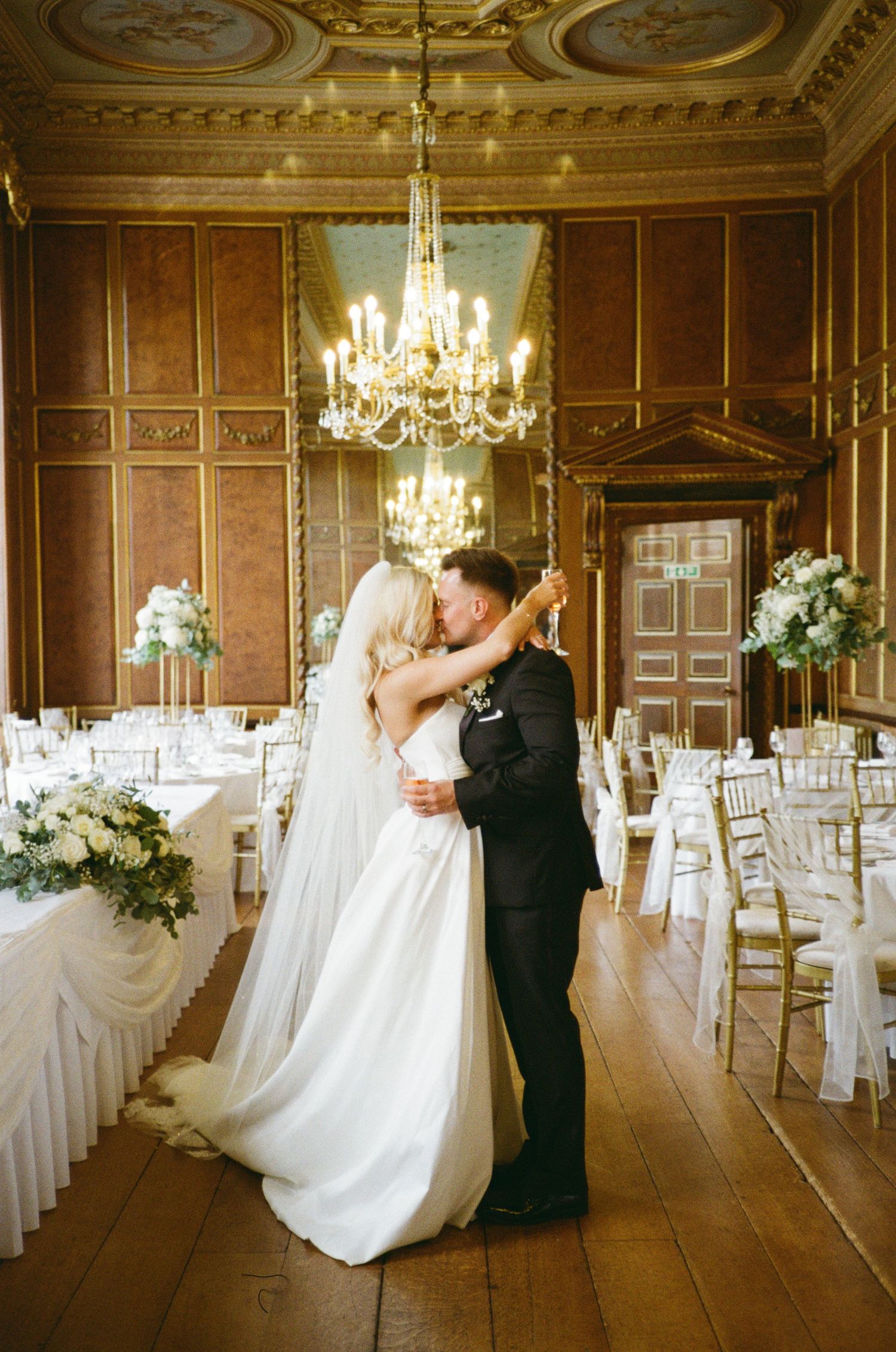 Romantic wedding couple dances in an elegant ballroom with ornate ceiling, chandeliers and white-draped chairs.
