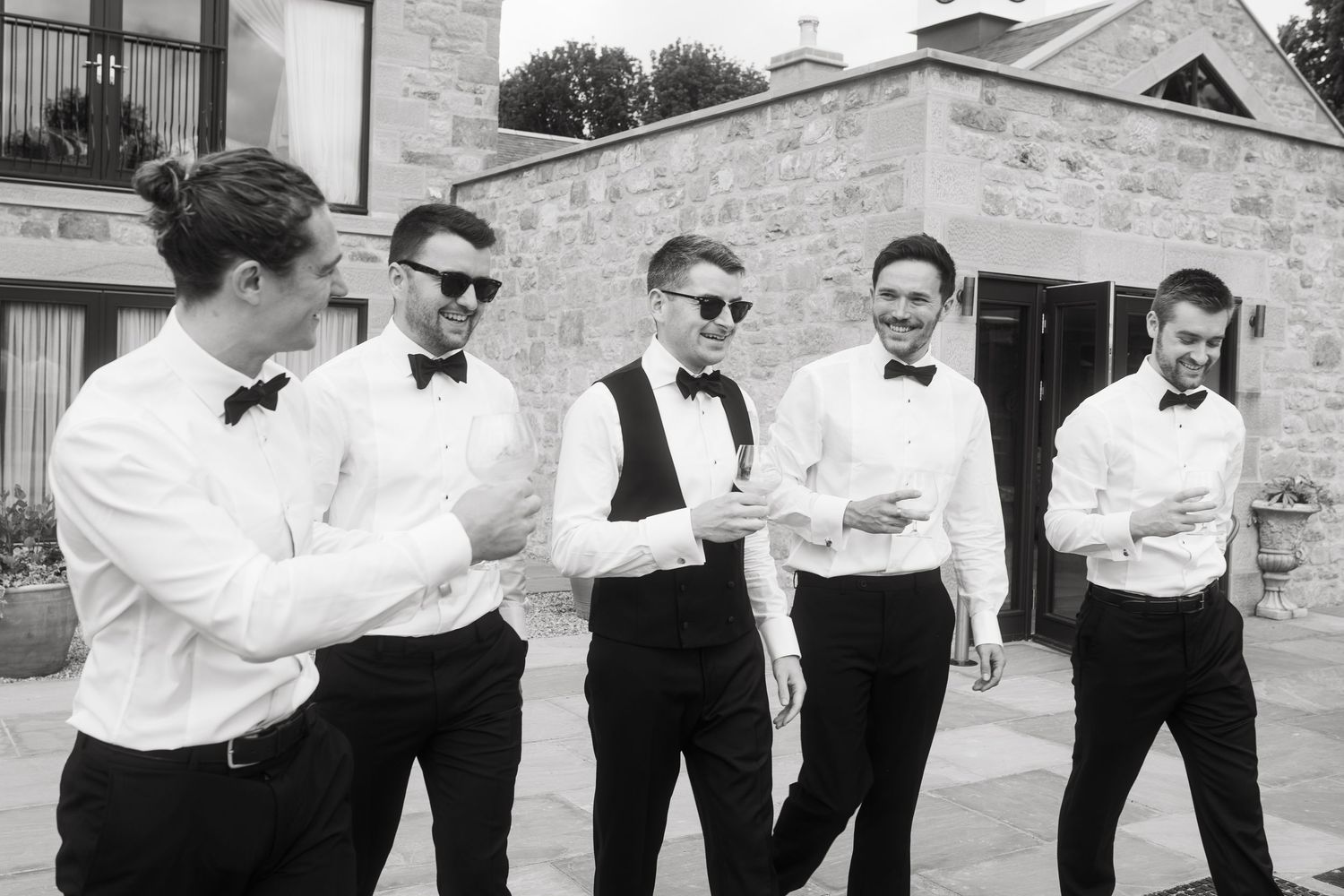 Groomsmen in black suits and bowties stand in row making funny poses at outdoor wedding venue.