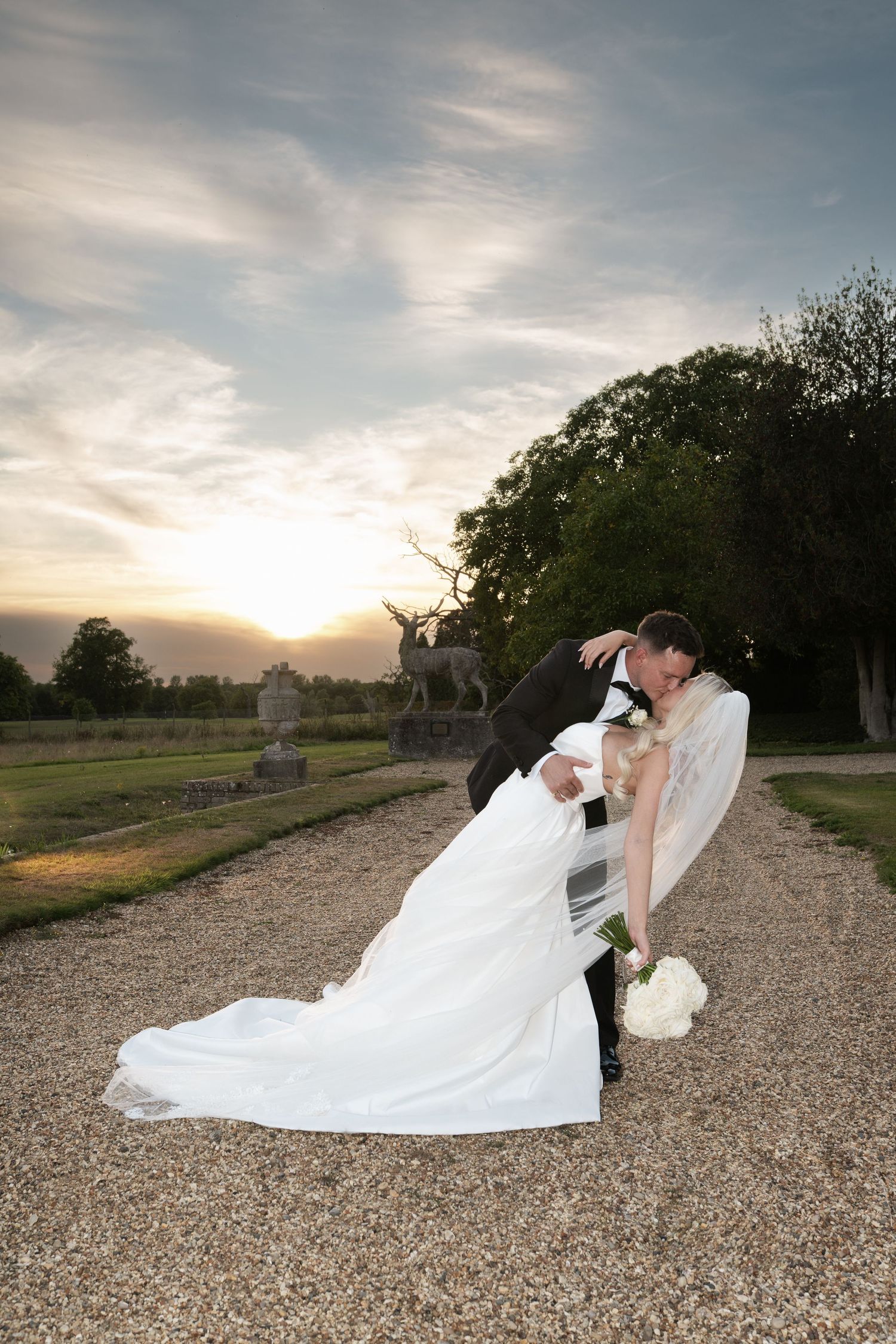 A romantic couple in formal attire share an intimate moment at sunset on a gravel path near trees.