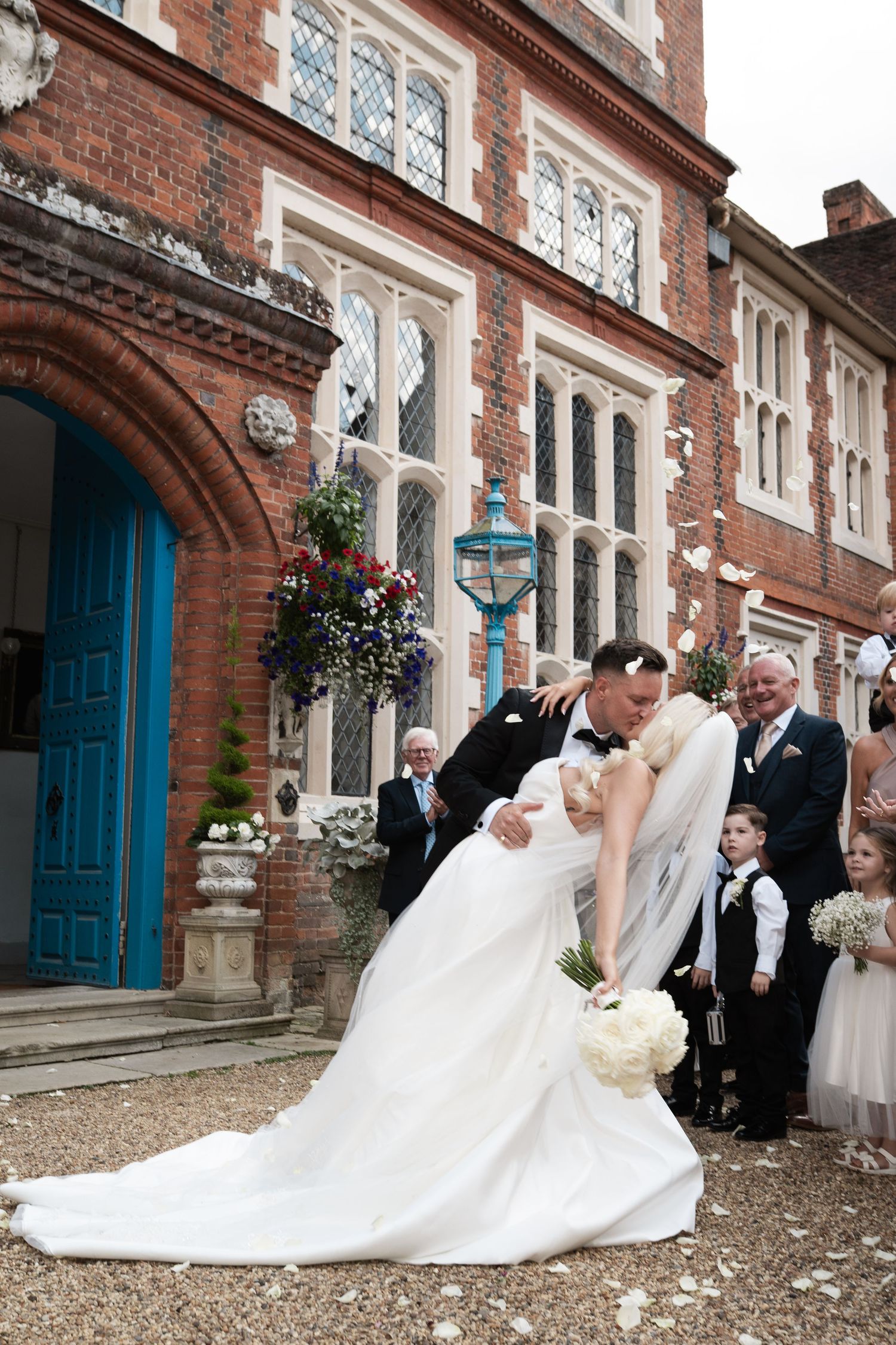Wedding party gathers outside historic brick building with elegant white gowns and formal attire for traditional photos.