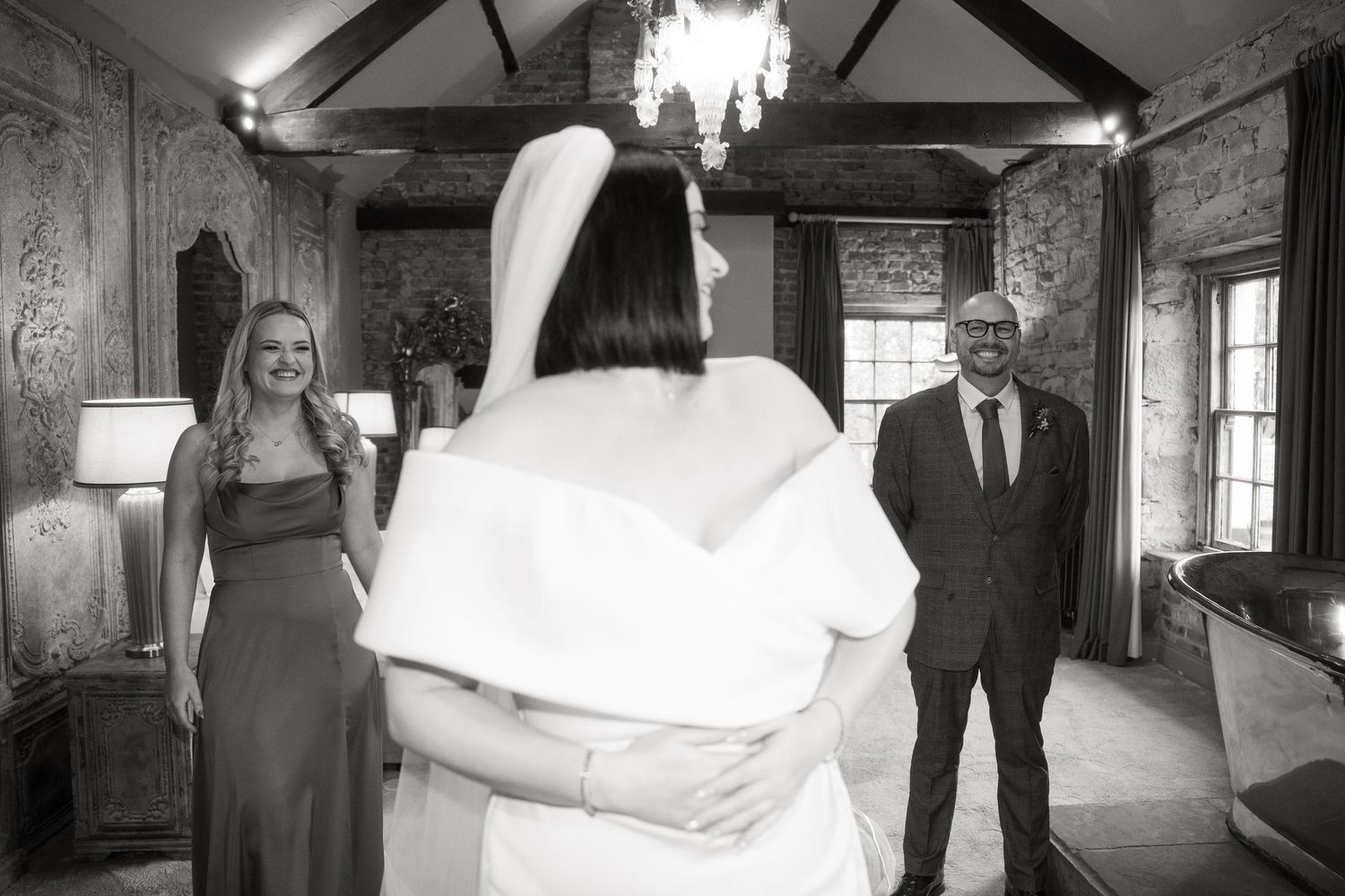 A black and white photo of a wedding ceremony moment in a rustic room with exposed beams and a chandelier.