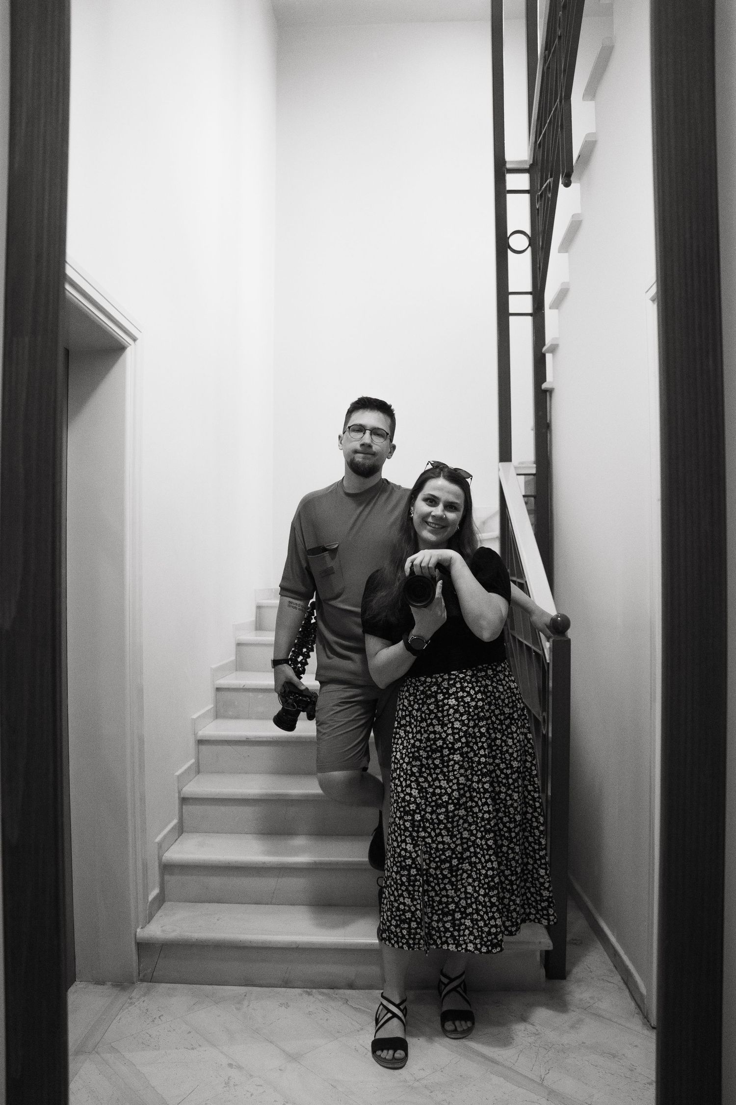 A black and white photograph of a couple standing together on a staircase in a narrow hallway.