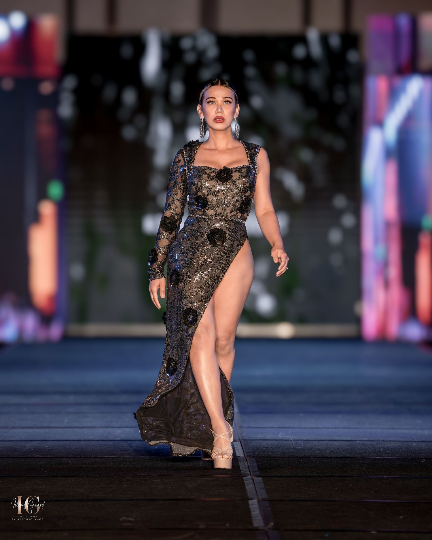 Model walks runway in black lace evening gown with high slit and sheer sleeves at fashion show.