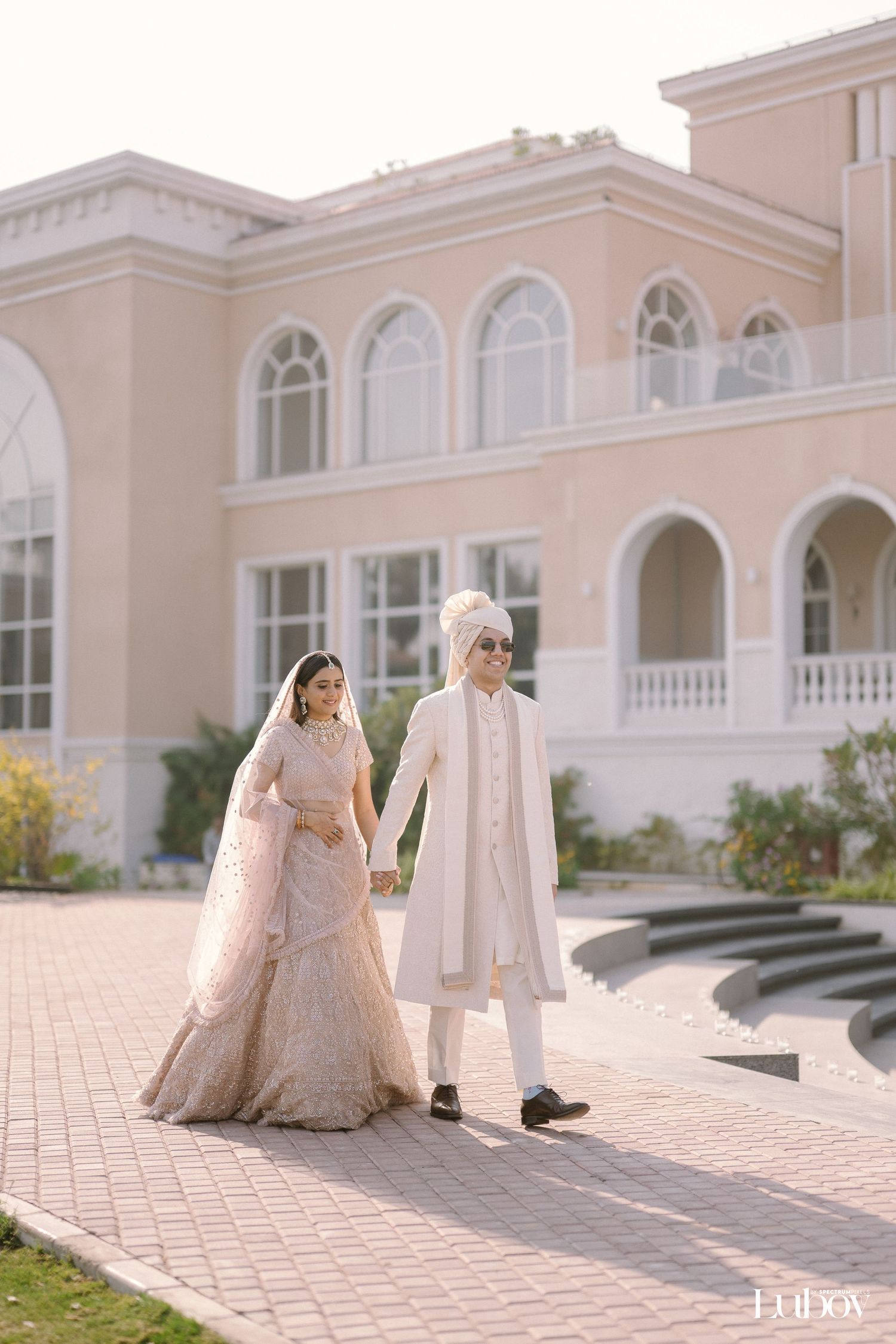 A couple in elegant blush-toned wedding attire stand holding hands outside a grand pink mansion with arched windows.