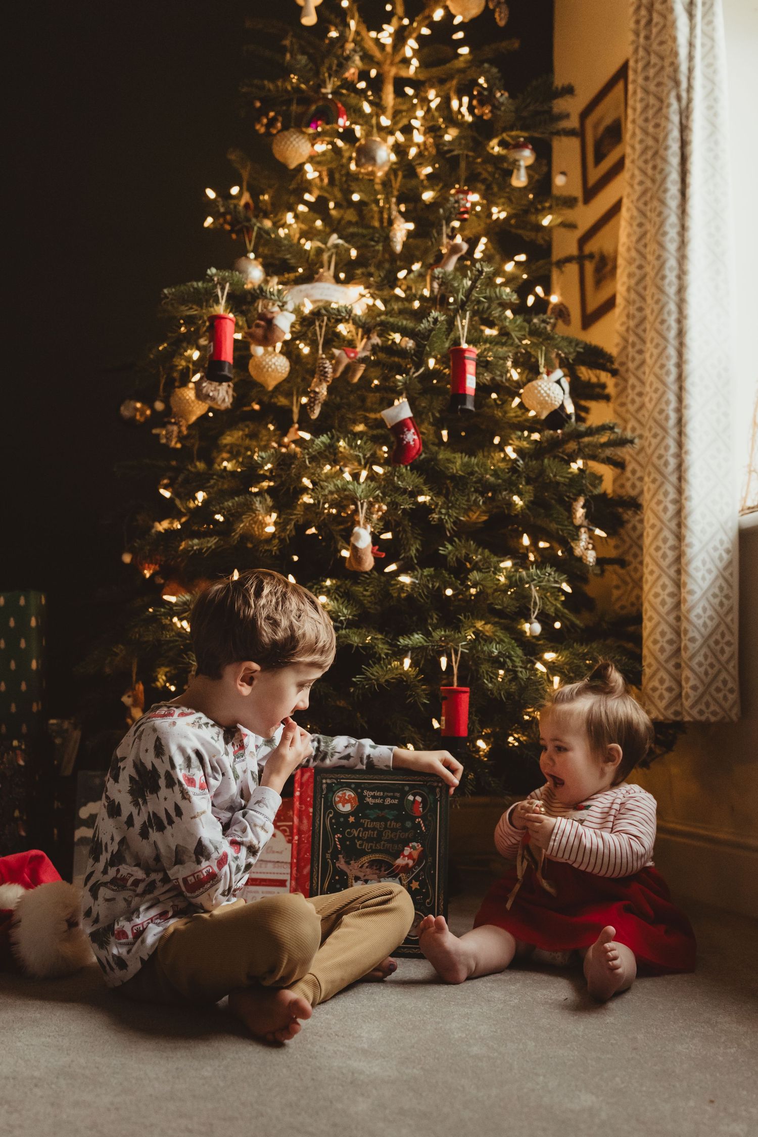 Children in matching holiday pajamas lay on the floor looking at Christmas presents under a decorated tree.