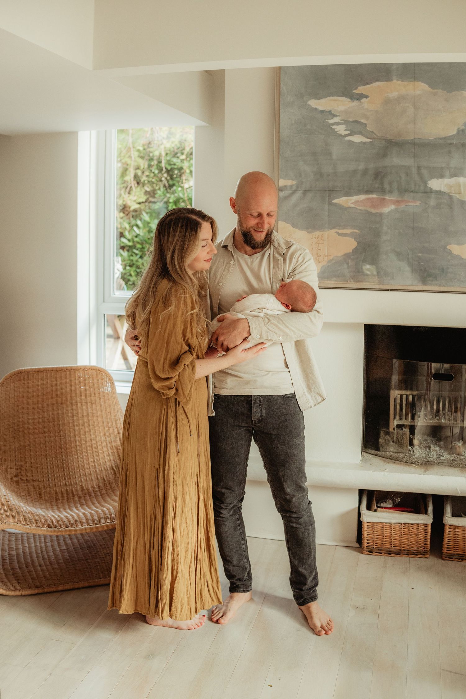 A couple stands together in a cozy living room with a fireplace, holding a newborn baby near a modern rattan chair.