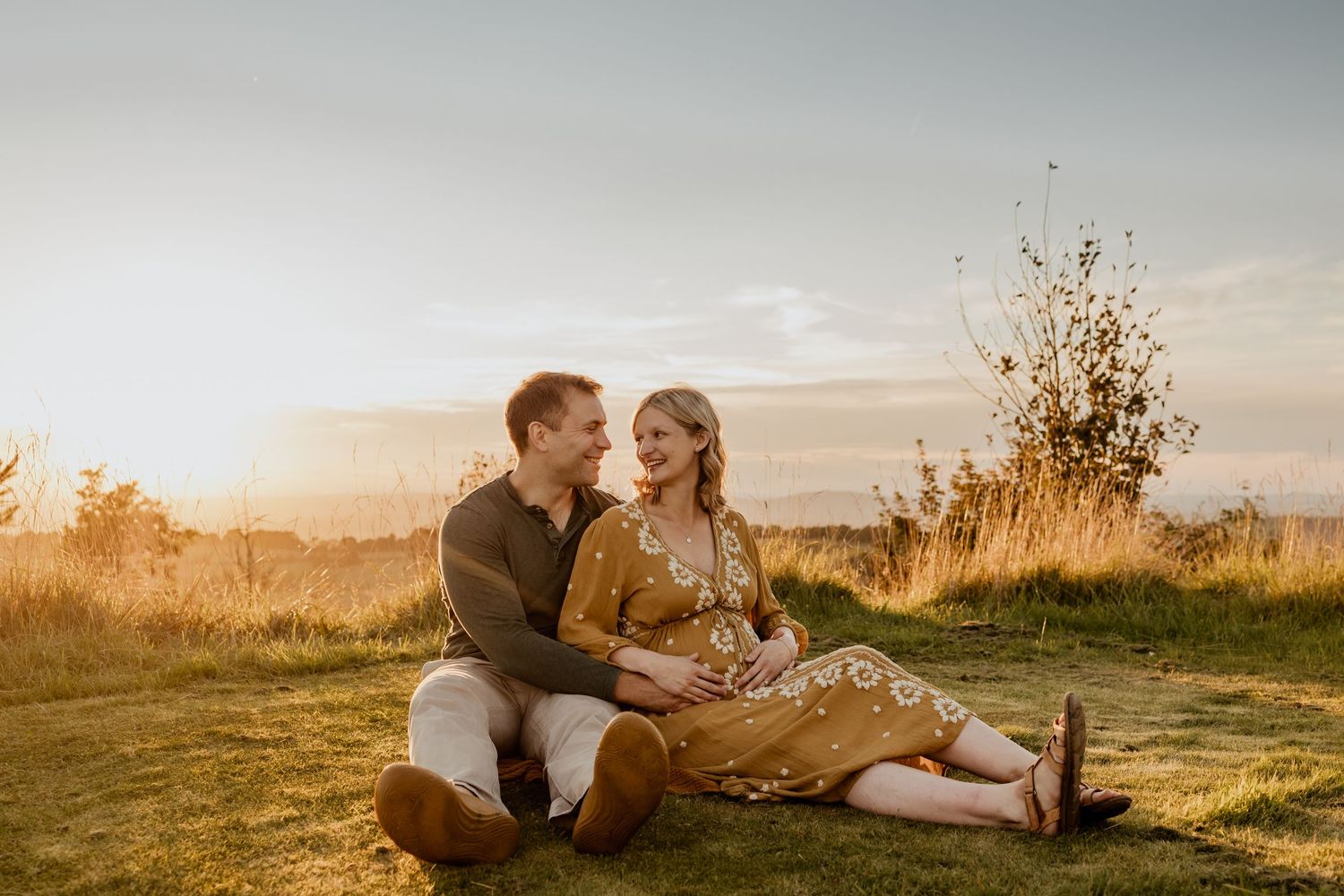 Romantic couple lying in golden sunlit meadow during sunset while taking selfies together on their phones.