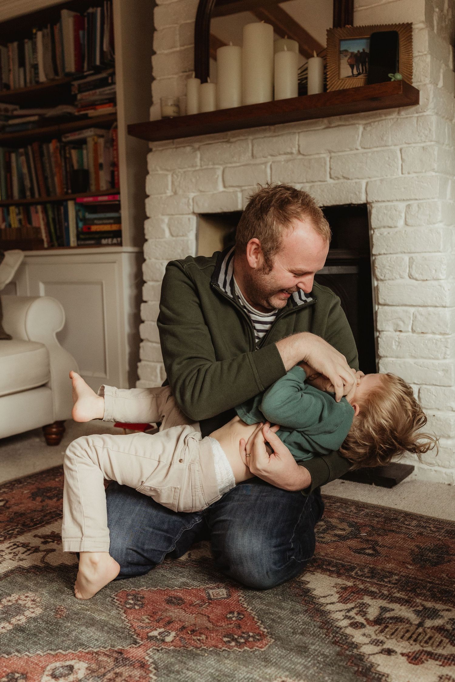 A parent and child share a playful moment on a patterned rug in a cozy room with bookshelves and white brick walls.