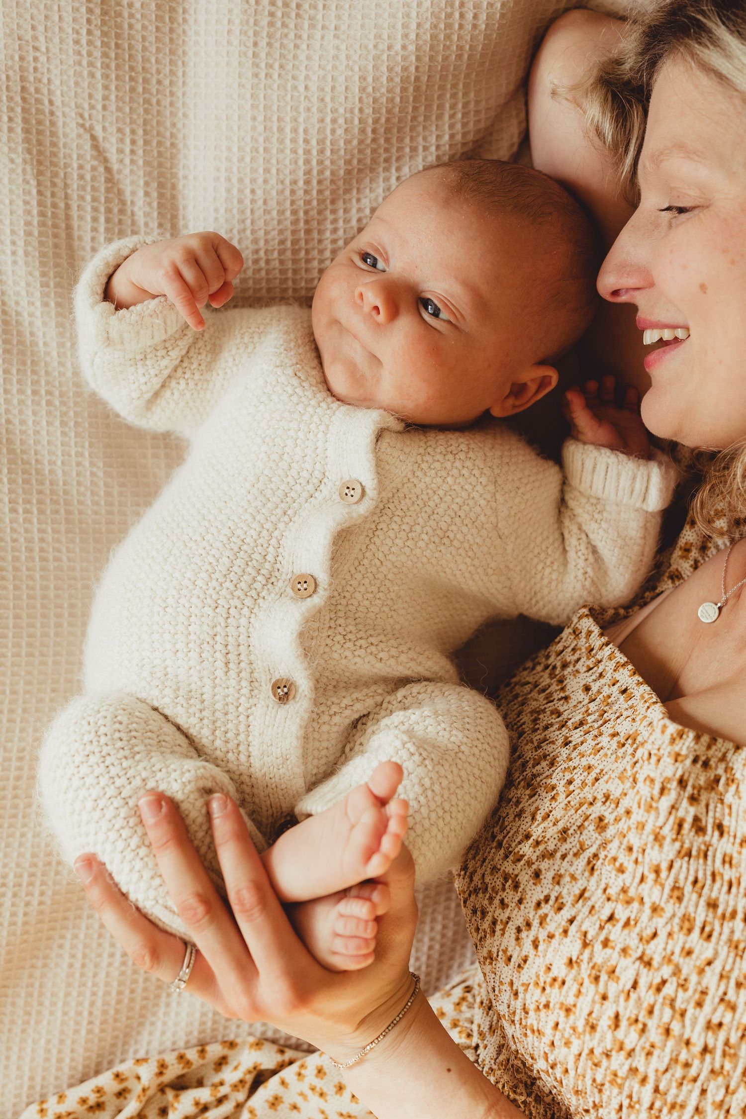 A tender moment between parent and baby in soft cream knitted sweaters laying on white bedding.