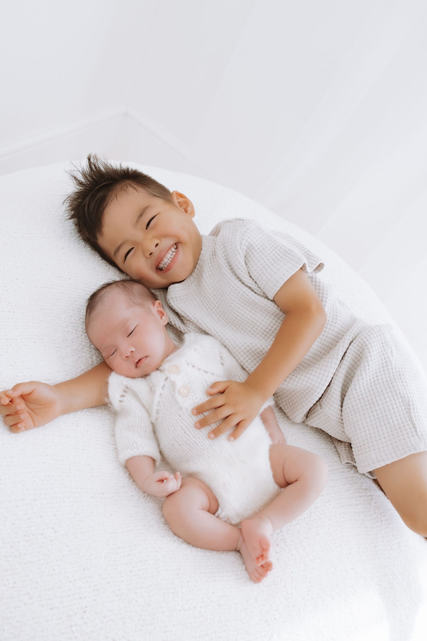 Two young siblings lying together on white bedding while wearing light colored clothing and smiling.