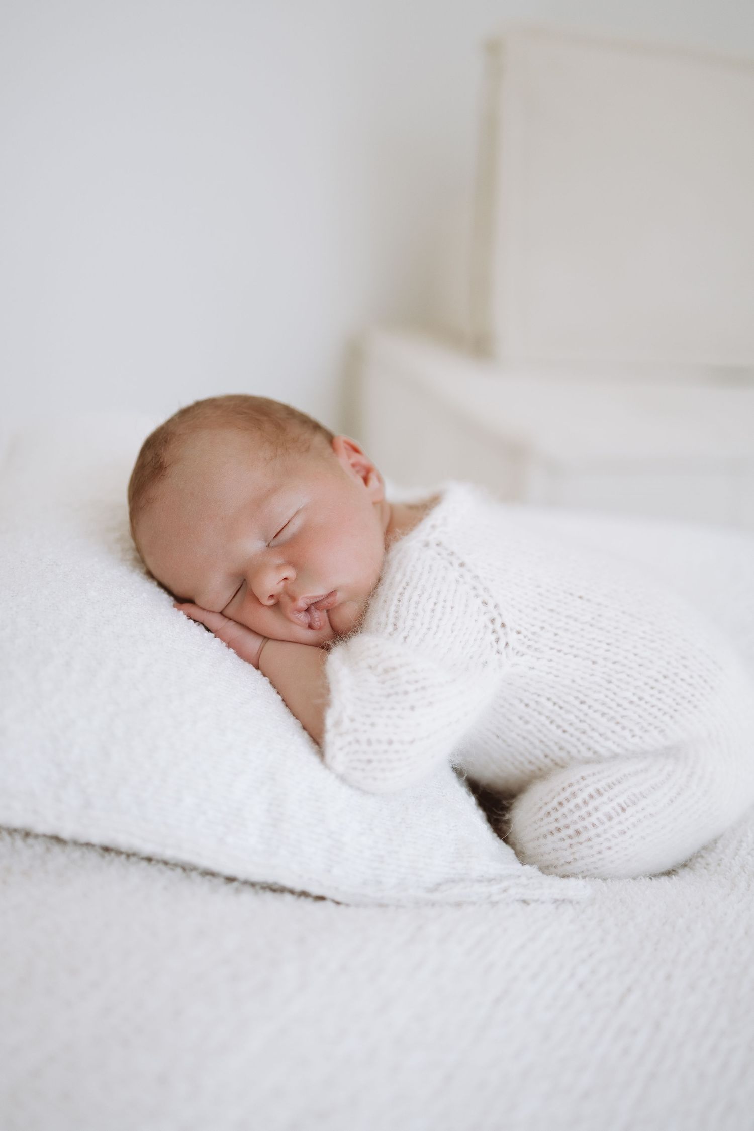Sleeping newborn baby wrapped in soft white knit blanket against white textured background.