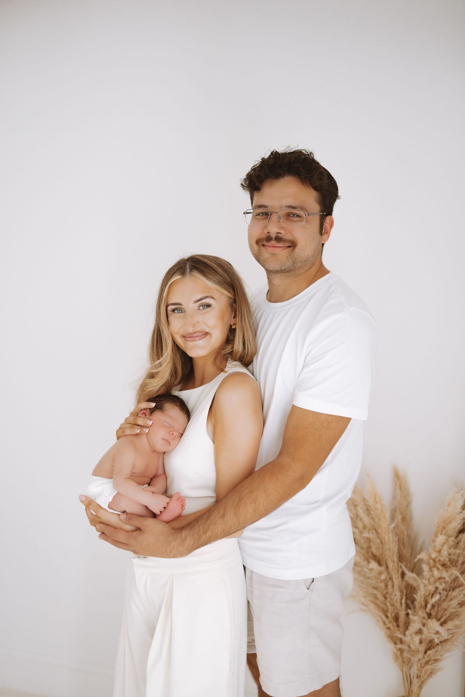 A couple in white outfits embraces while holding a newborn baby, with pampas grass decor in the background.