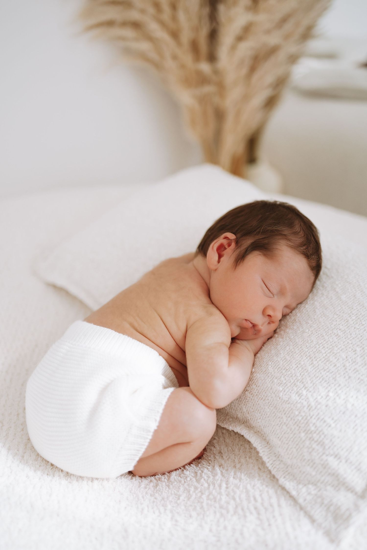 A sleeping newborn baby rests peacefully on soft white textiles in natural window light from multiple angles.