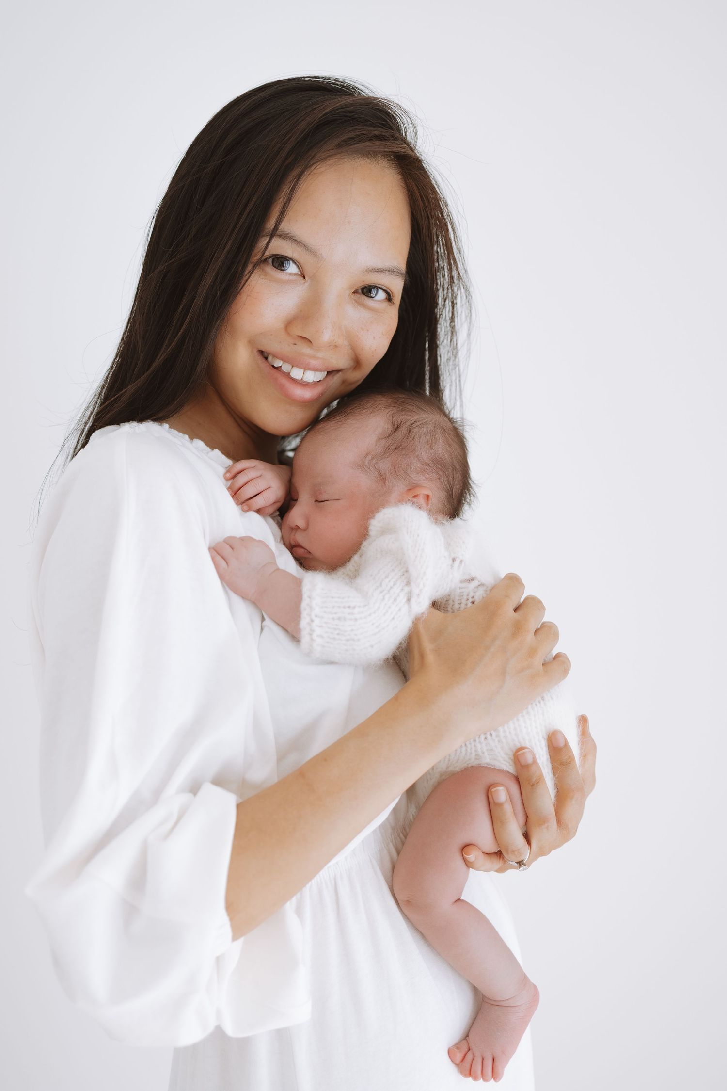 Parent in white dress holding sleeping newborn baby against white background.