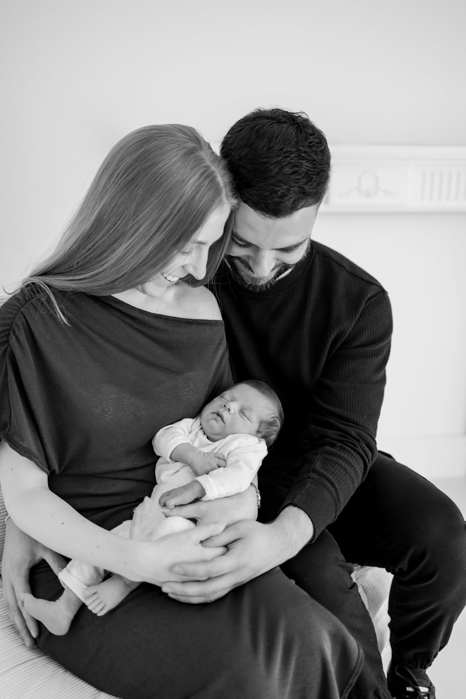 Two people in black clothing hold and gaze lovingly at a newborn baby in a monochrome portrait series.