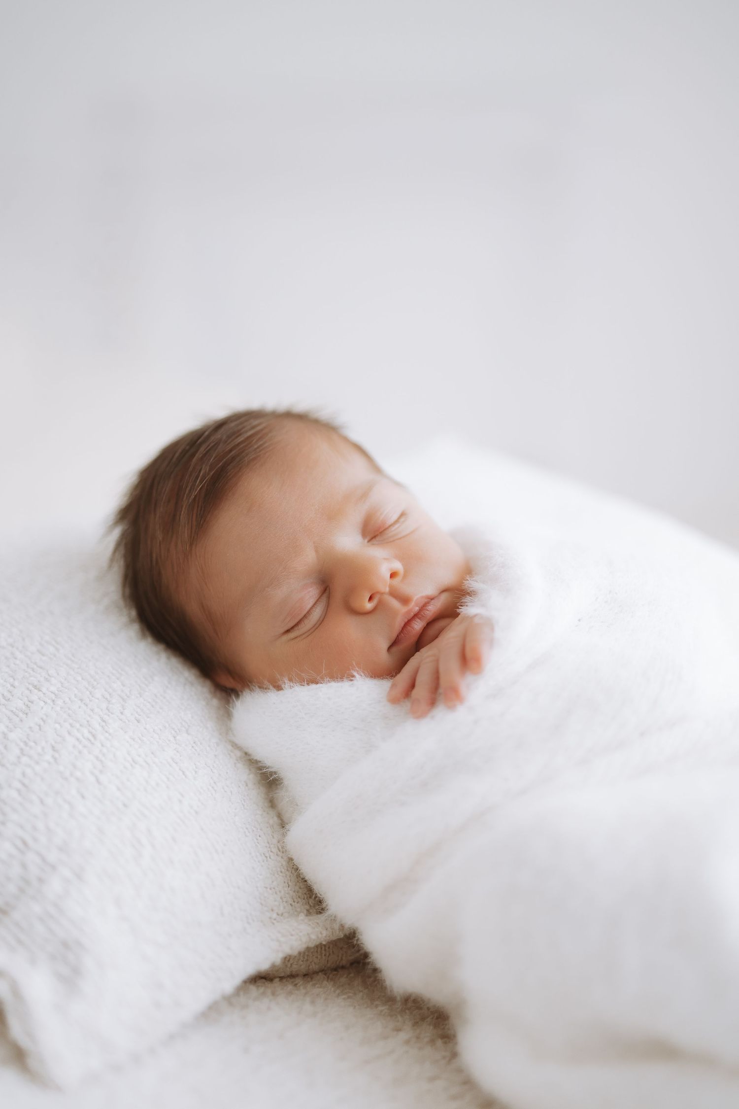 A sleeping newborn baby wrapped in soft white swaddle blankets photographed in a dreamy, minimalist style.