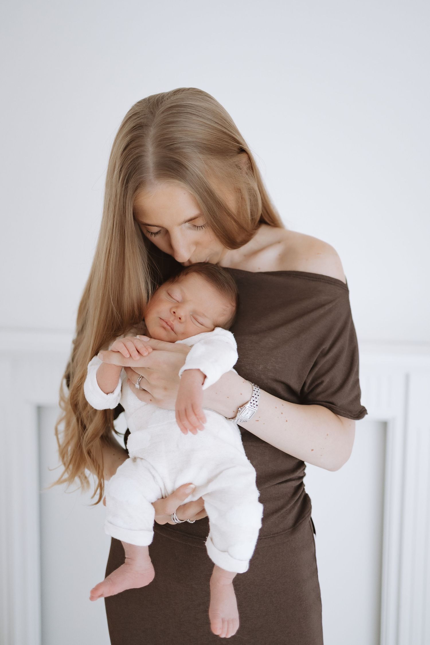 A parent in a brown sweater tenderly holds and kisses a baby dressed in white.