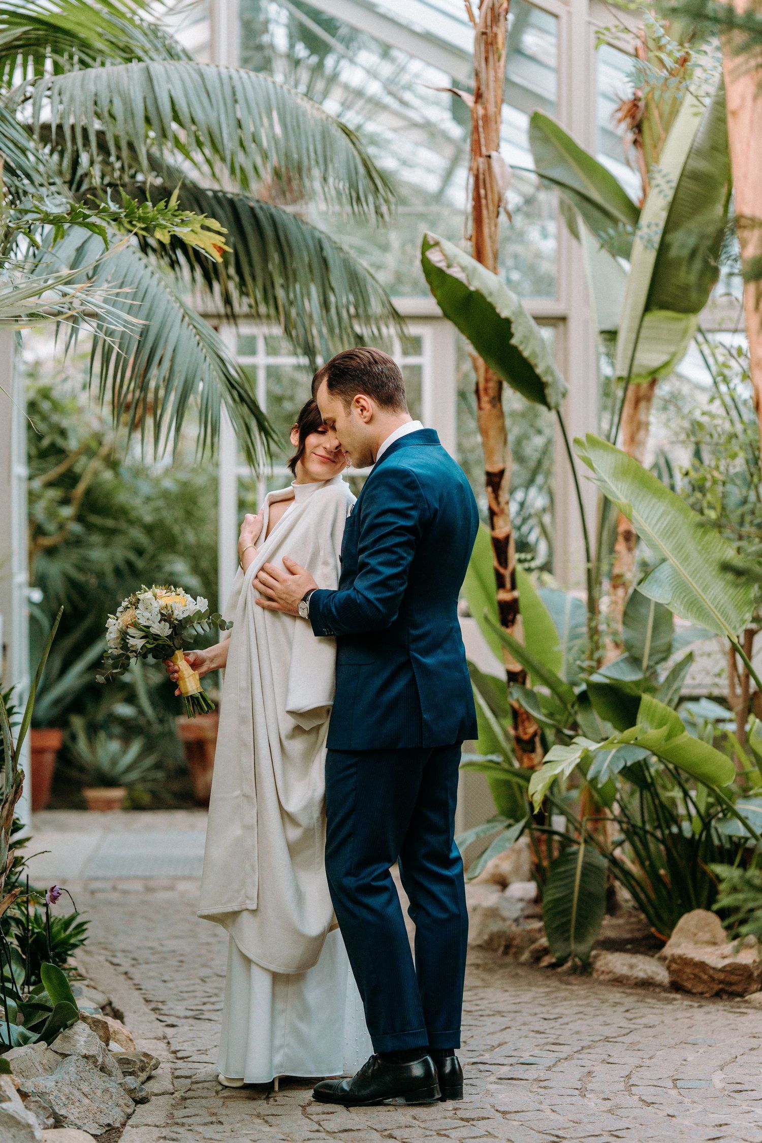 A romantic couple embraces in a lush greenhouse setting with tropical palms and banana plants surrounding them.