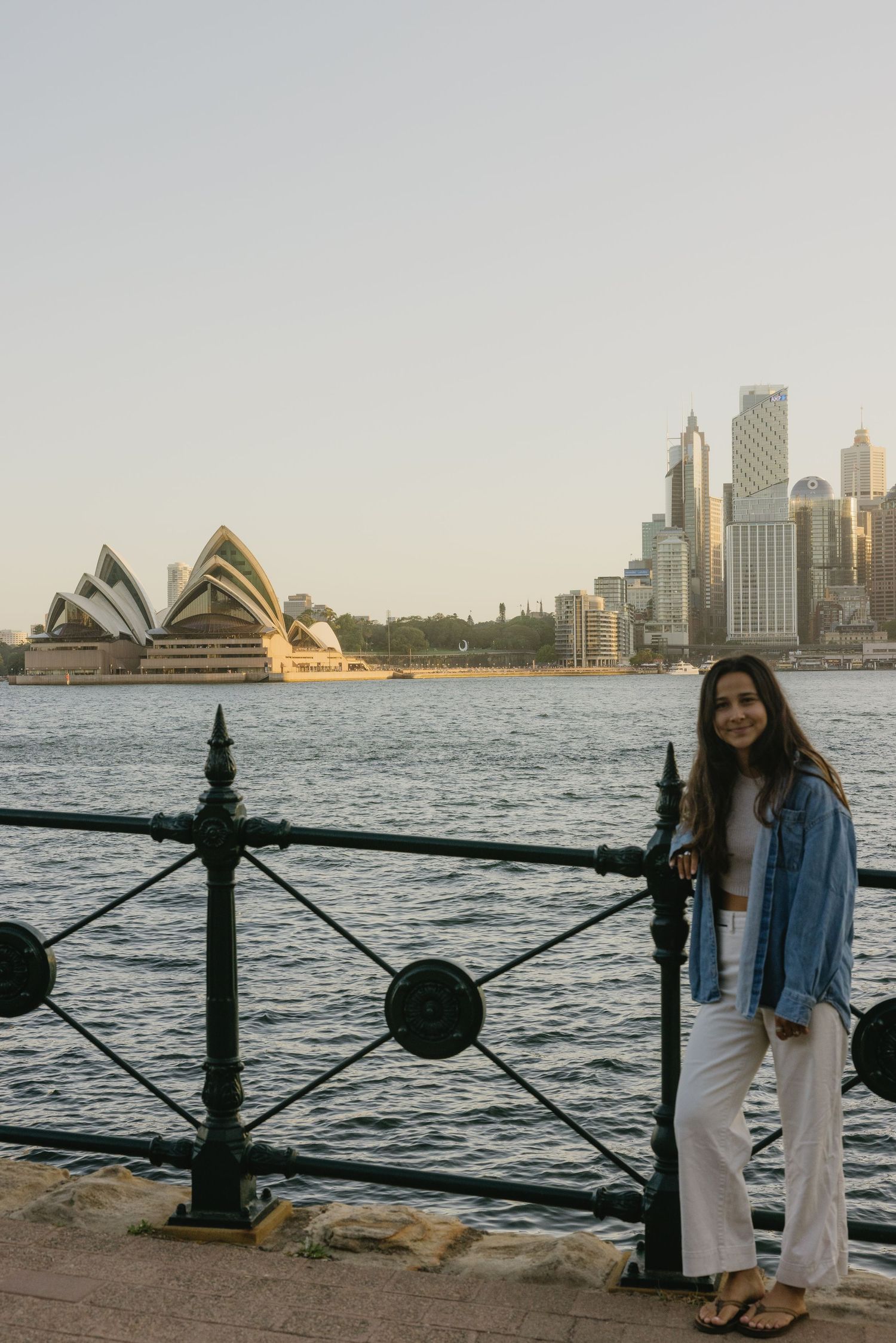 Woman posing by Sydney Harbour railing with iconic Opera House and city skyline in background at sunset.