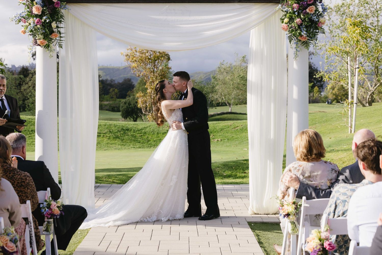 A couple shares their first kiss under an elegant outdoor wedding arch decorated with floral arrangements.