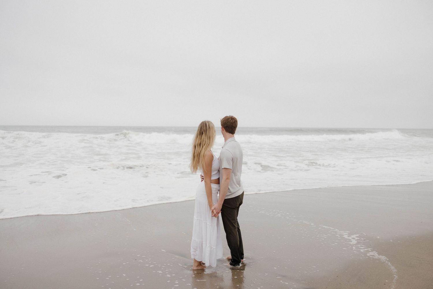 A couple stands together on a misty beach with waves rolling in as the sea meets the sand at the shoreline.