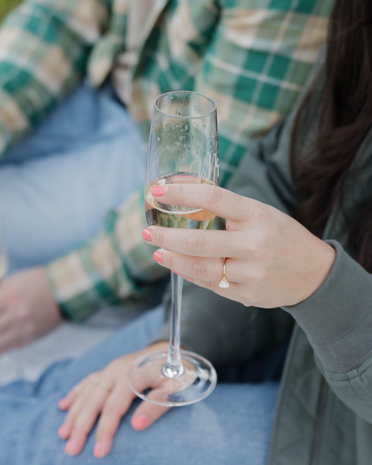 A hand holding a champagne flute with a ring visible on the finger against a blurred plaid background.