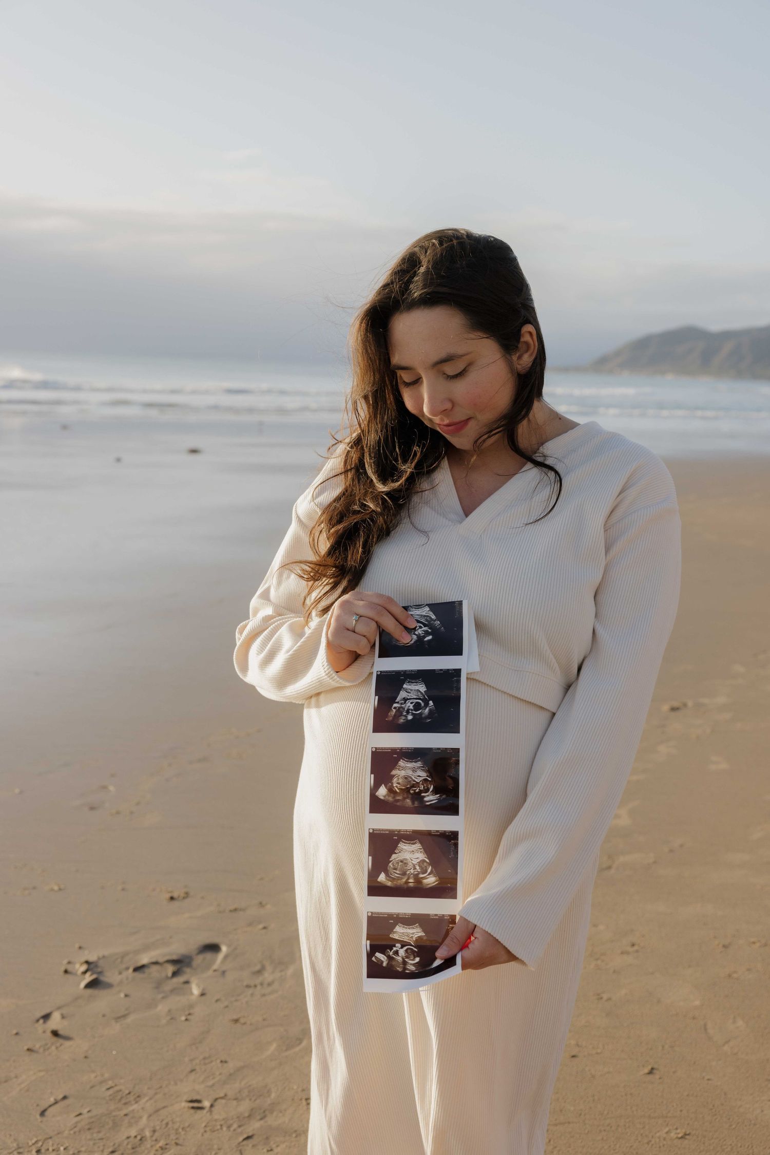 Person in cream-colored dress holds ultrasound photos while standing on beach at sunset.