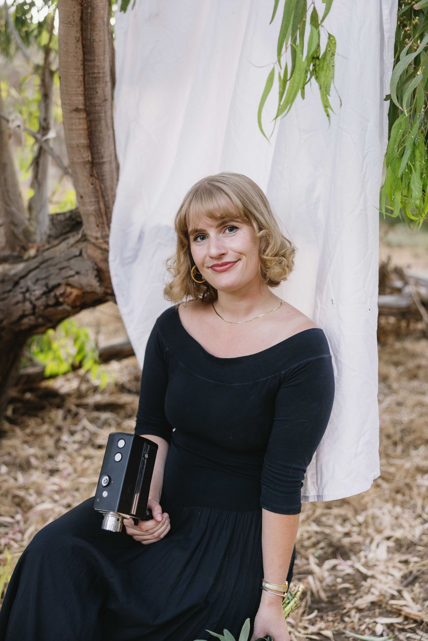 Someone in a black dress sits outdoors against a white backdrop while holding a vintage camera and smiling.