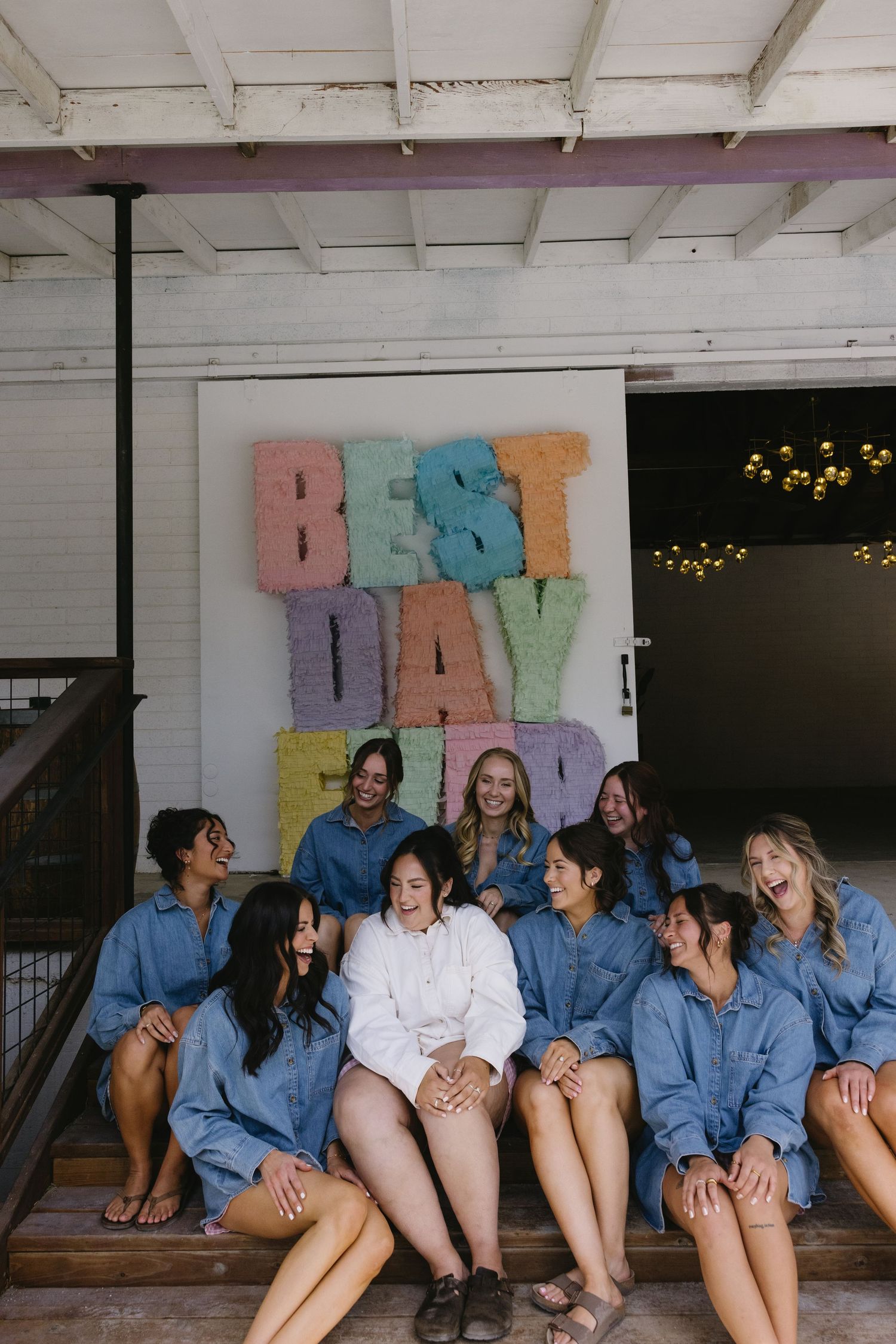 Bridal party in matching denim shirts laughing on steps in front of a colorful Best Day Ever sign.