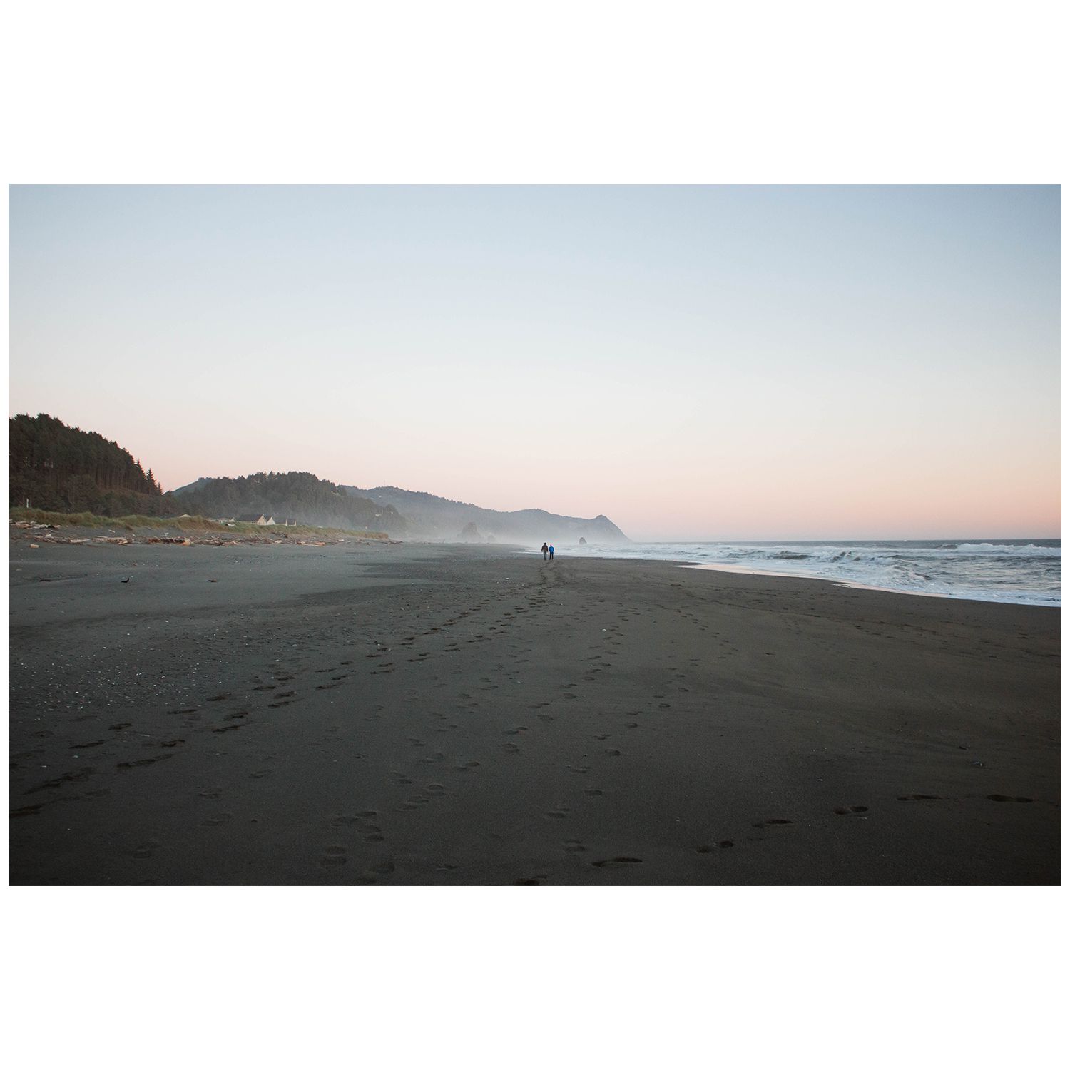 Silhouettes walk along a misty beach at dusk with coastal cliffs and rolling waves visible in the distance.