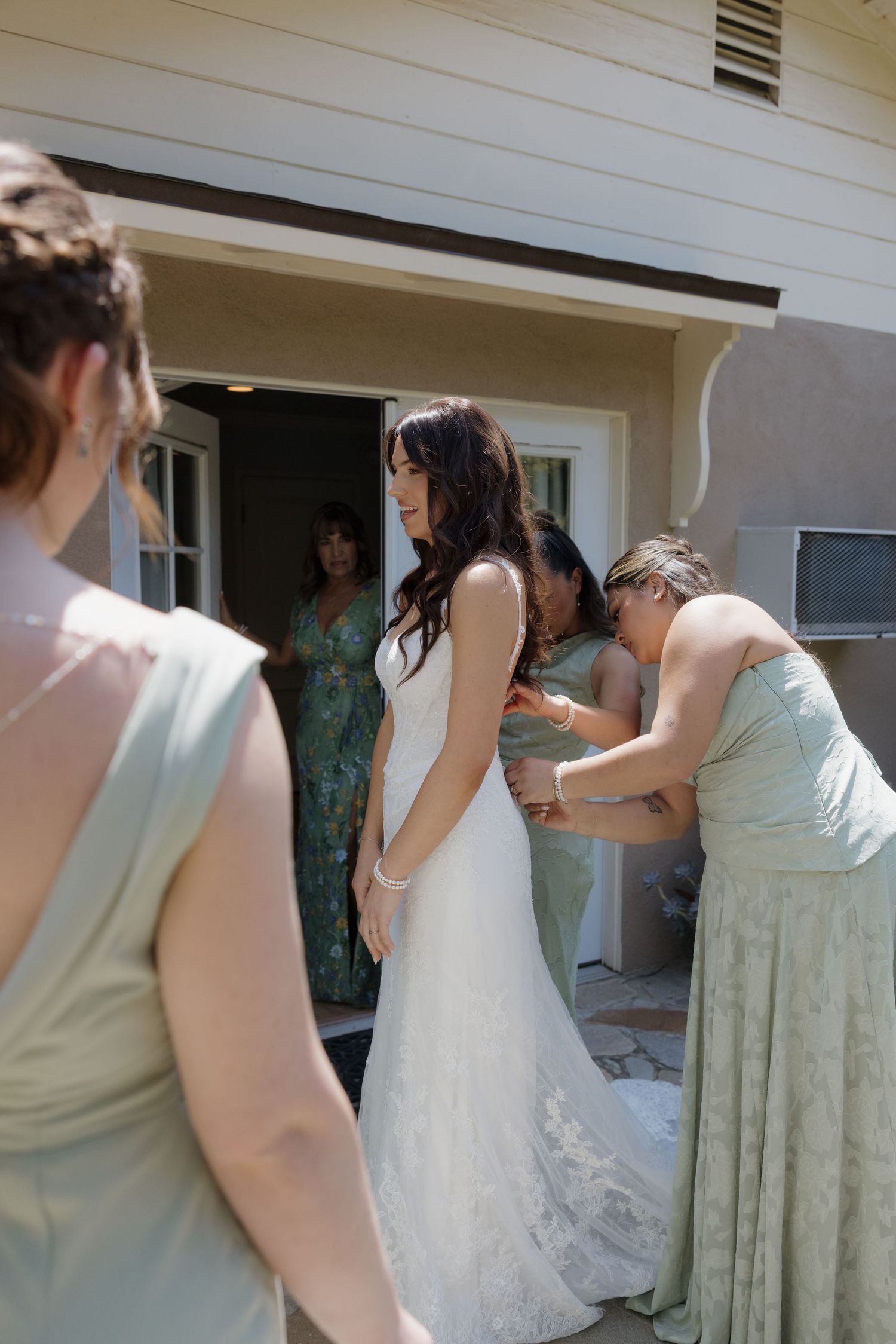 Bride in white lace gown being helped by bridesmaids in sage green dresses outside a home before wedding ceremony.