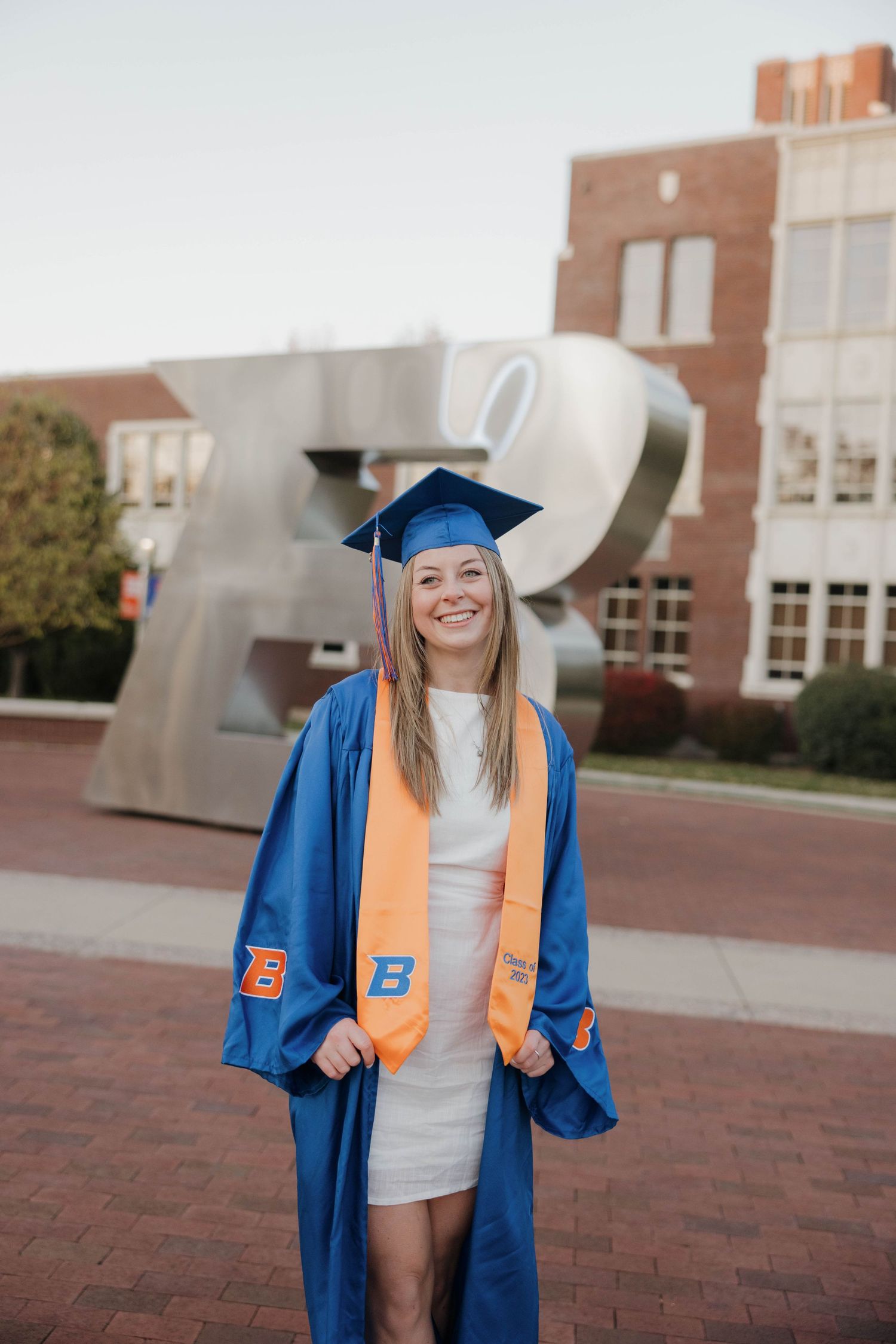 Graduate in blue cap and gown with orange stole stands on brick plaza in front of modern university building sculpture.