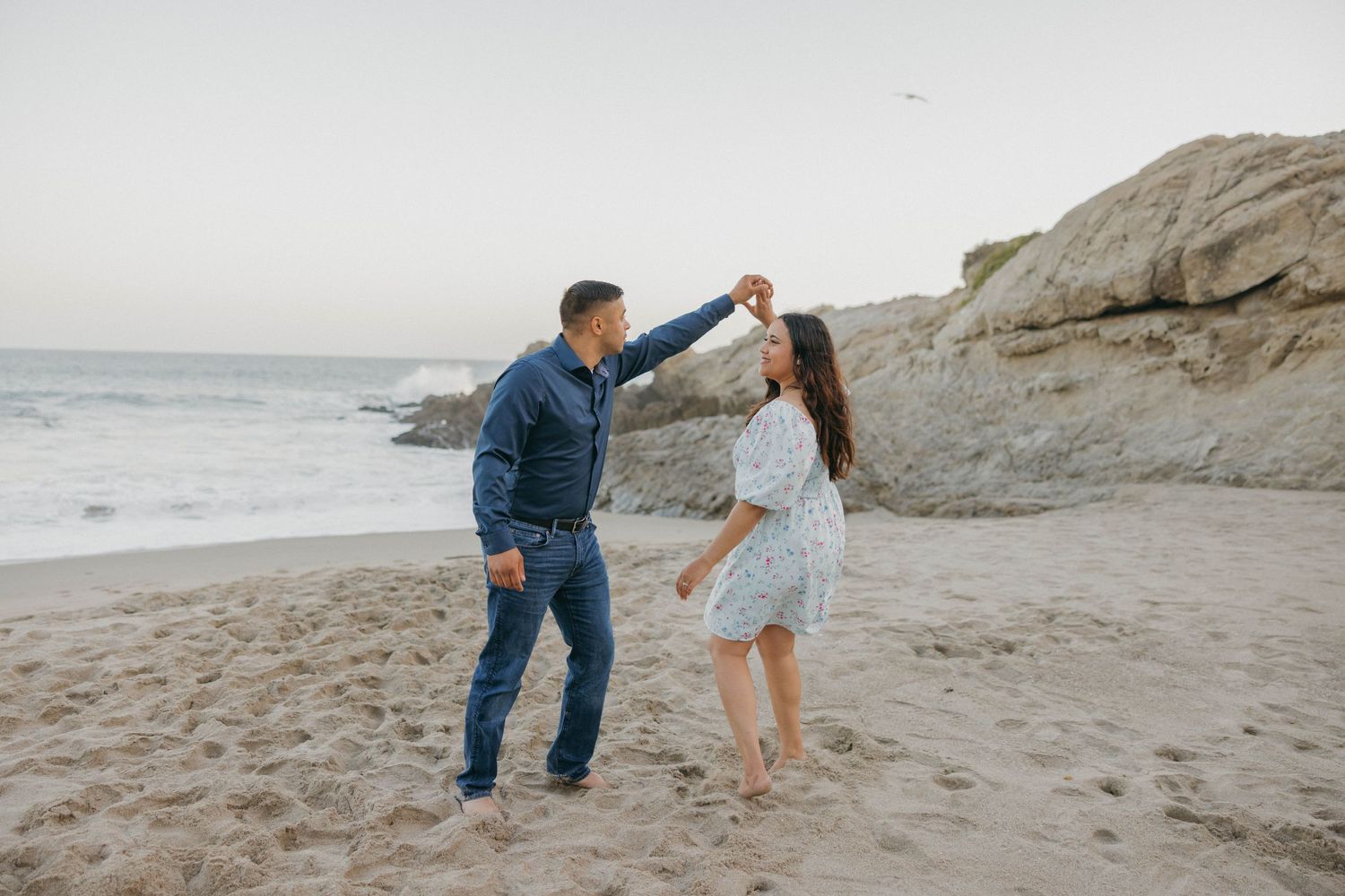 A couple dancing together on a sandy beach with rocky cliffs and ocean waves in the background.
