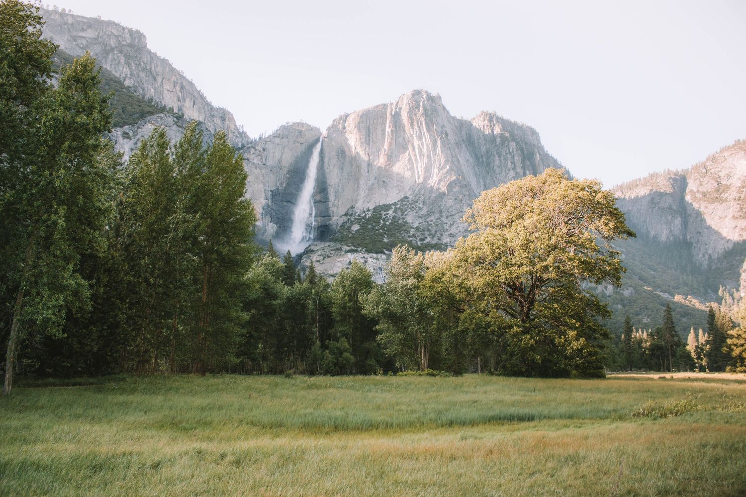 Majestic waterfall cascades down mountain cliffs in Yosemite Valley with green meadow in foreground.