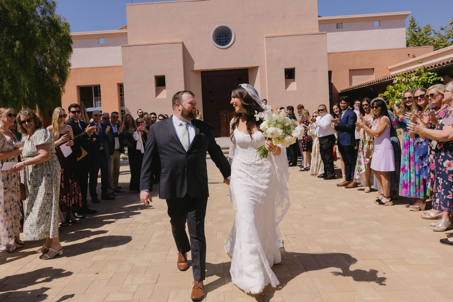 Bride and groom walk through cheering guests outside a sunlit venue after their wedding ceremony.
