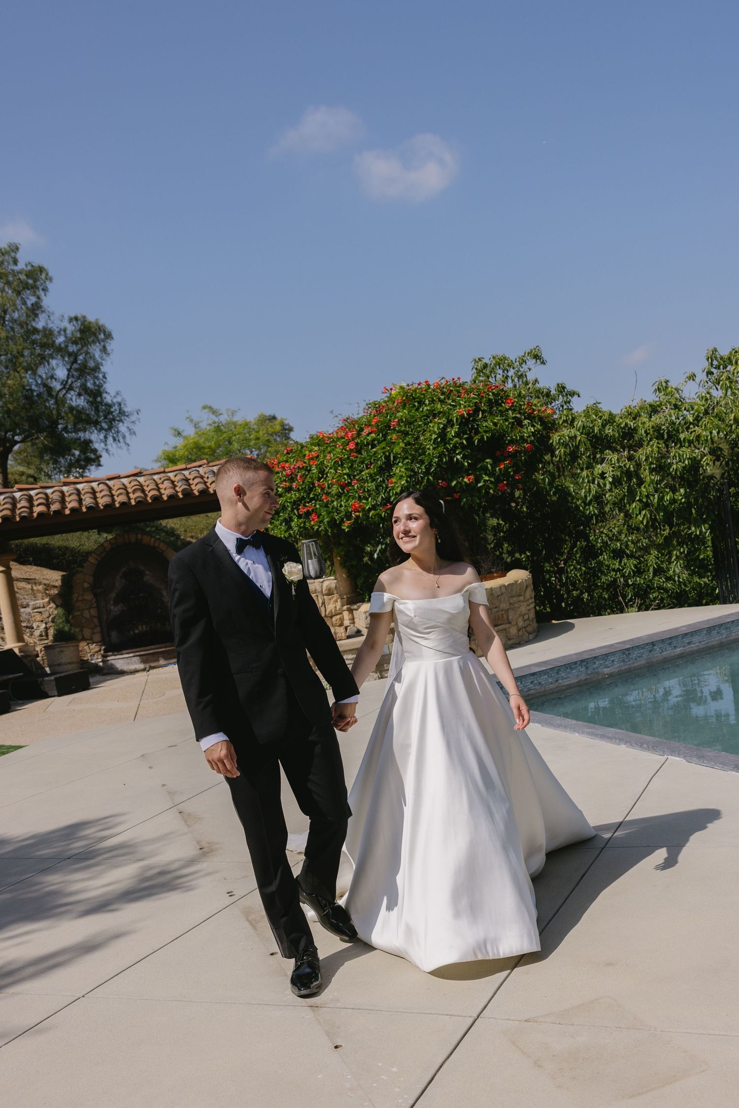 Elegant couple walking together beside a swimming pool at an outdoor venue on a sunny day with lush landscaping.