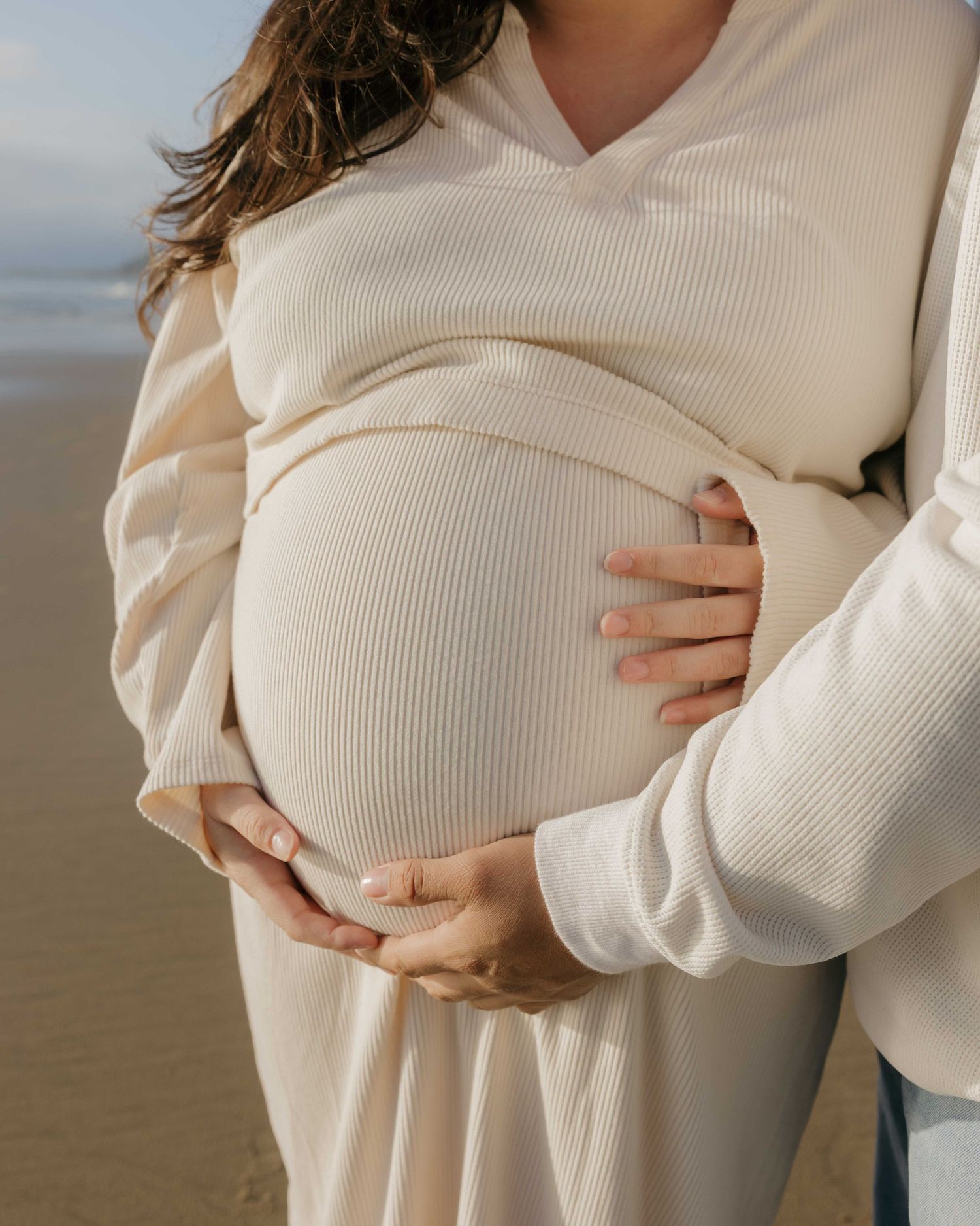 A pregnant woman in a cream ribbed sweater holds her belly tenderly while standing on a beach.